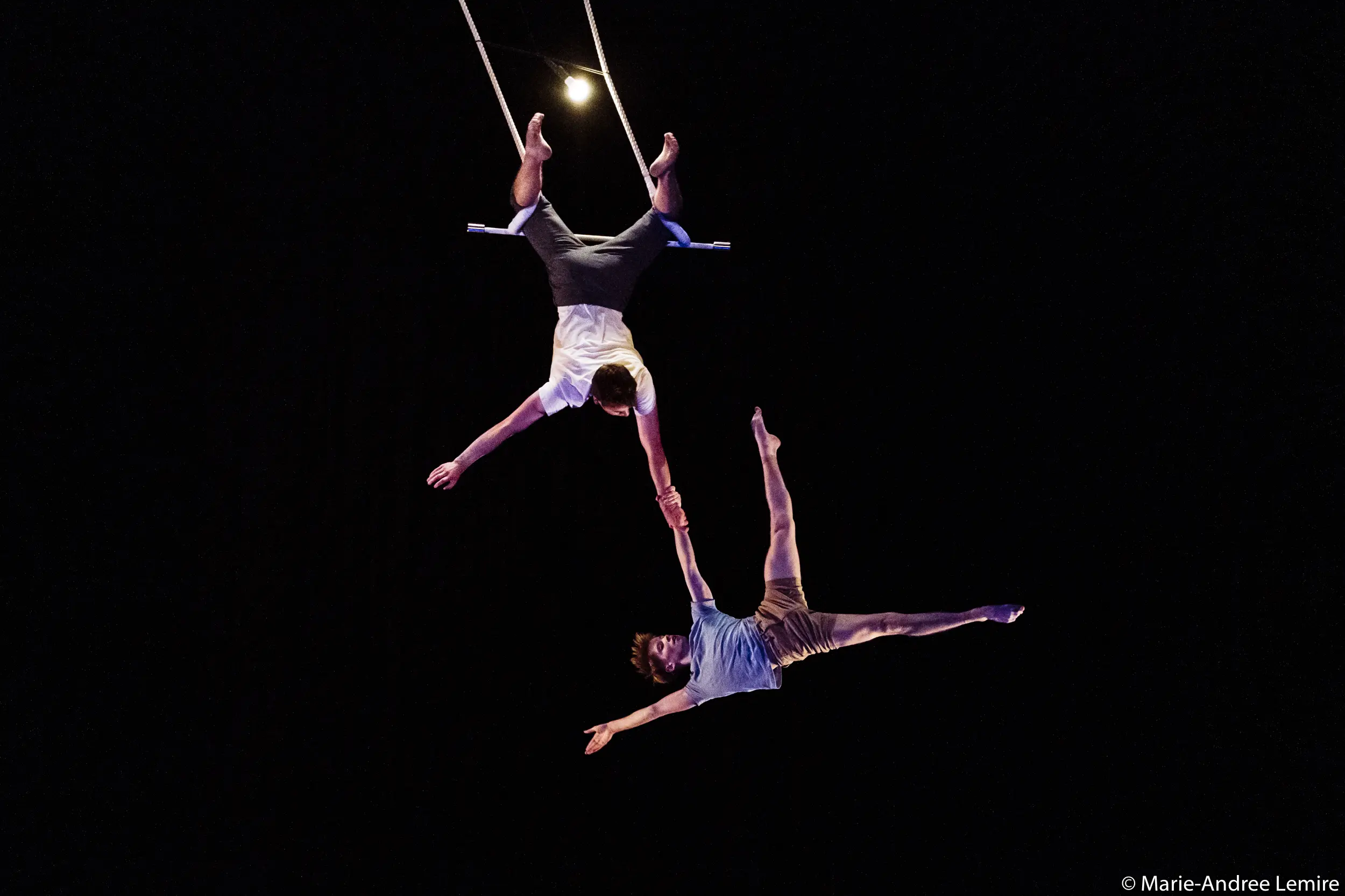 Deux acrobates, Maxime Charron et Corentin Lemaître Auger, sont suspendus en l'air sur un trapèze, l'un à l'envers et tenant la main de l'autre, faisant preuve d'un équilibre et d'une force remarquables sur un fond sombre.