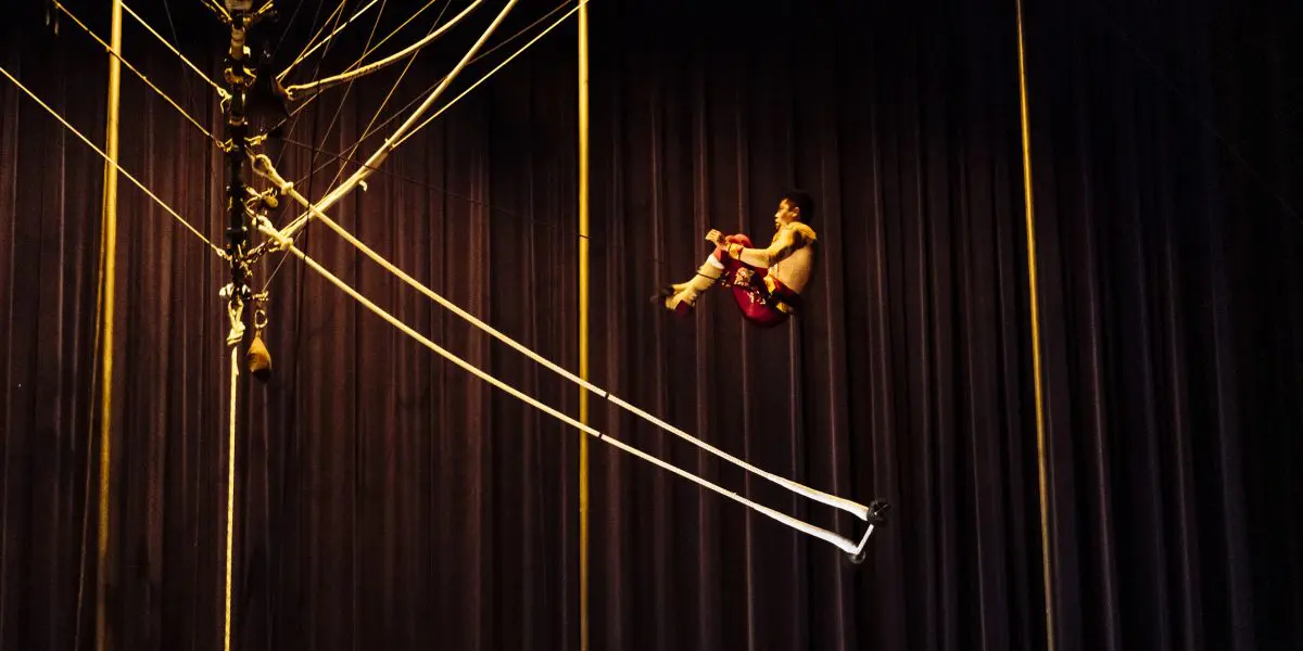 L'artiste de cirque Gabriel Olmos Cruz, en pantalon rouge, est suspendu à des cordes en hauteur contre un rideau bleu foncé. Le matériel de montage est visible sur le côté gauche de l'image.