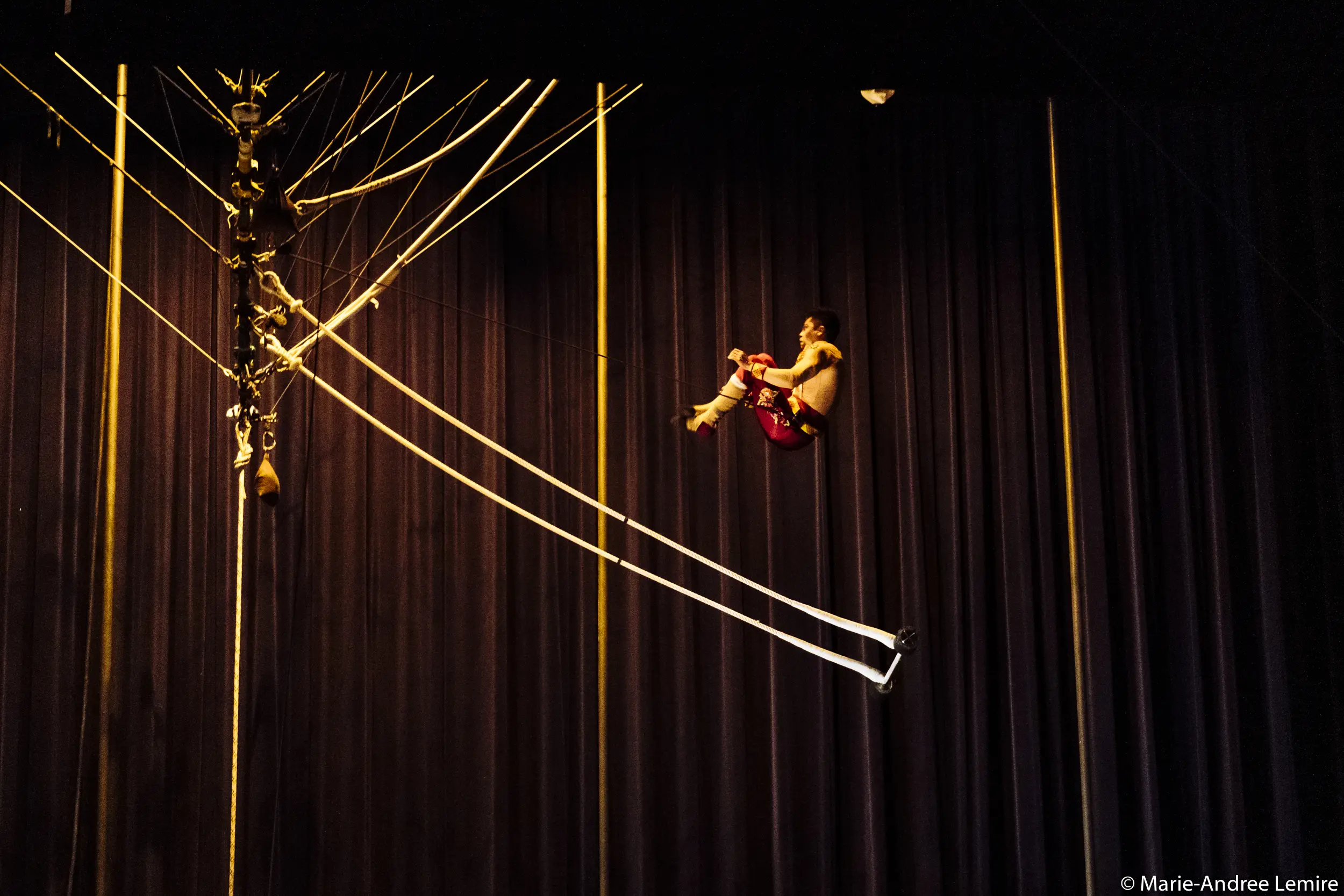 L'artiste de cirque Gabriel Olmos Cruz, en pantalon rouge, est suspendu à des cordes en hauteur contre un rideau bleu foncé. Le matériel de montage est visible sur le côté gauche de l'image.