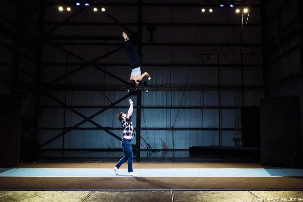 Une personne vêtue d'une chemise à carreaux se tient sur un tapis à l'intérieur d'un grand gymnase faiblement éclairé. Elle tend les bras vers le haut tandis qu'Ashley Youren s'élève au-dessus d'elle, en plein vol et la tête en bas, dans une pose acrobatique spectaculaire.
