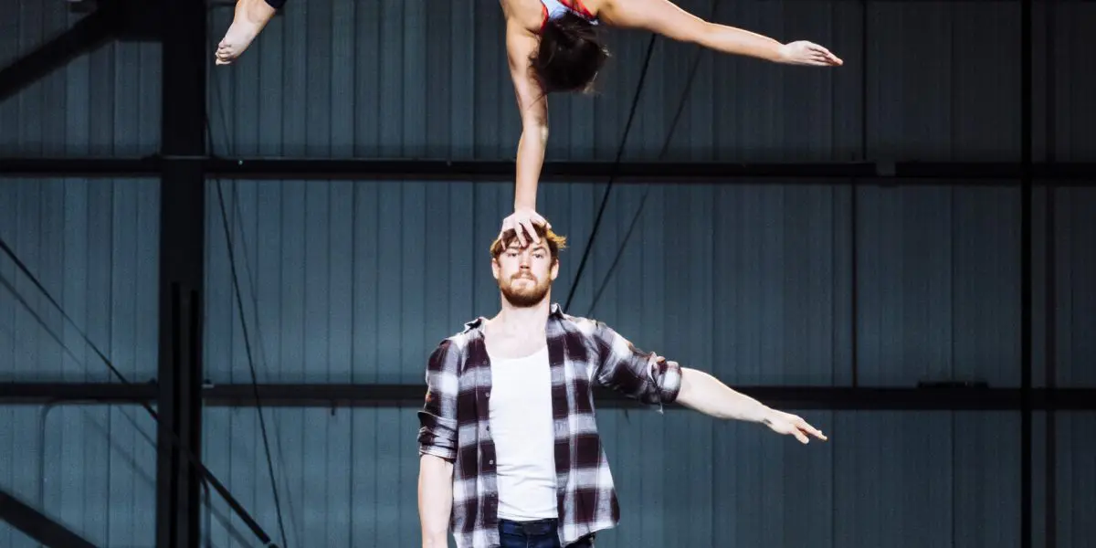 Un homme se tient debout sur un tapis, faisant tenir Ashley Youren à l'envers sur une main. Ses jambes forment une fente tandis qu'elle se soutient avec un bras tendu vers lui. Ils s'exécutent avec précision sur une scène intérieure.