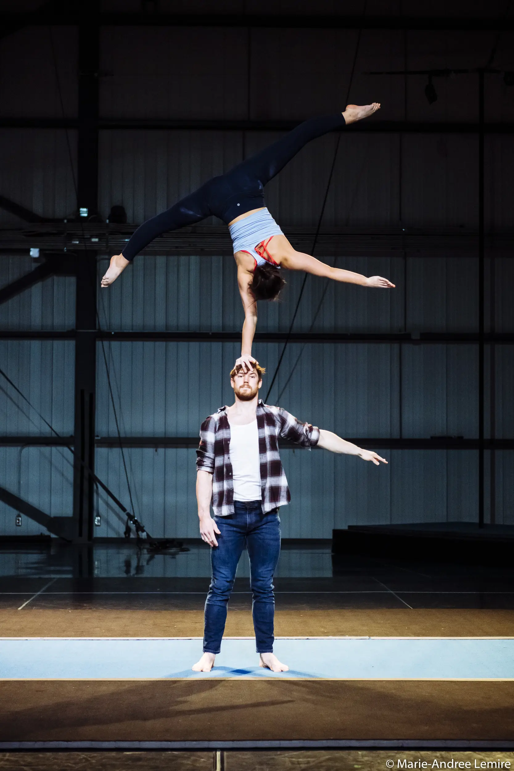 Un homme se tient debout sur un tapis, faisant tenir Ashley Youren à l'envers sur une main. Ses jambes forment une fente tandis qu'elle se soutient avec un bras tendu vers lui. Ils s'exécutent avec précision sur une scène intérieure.