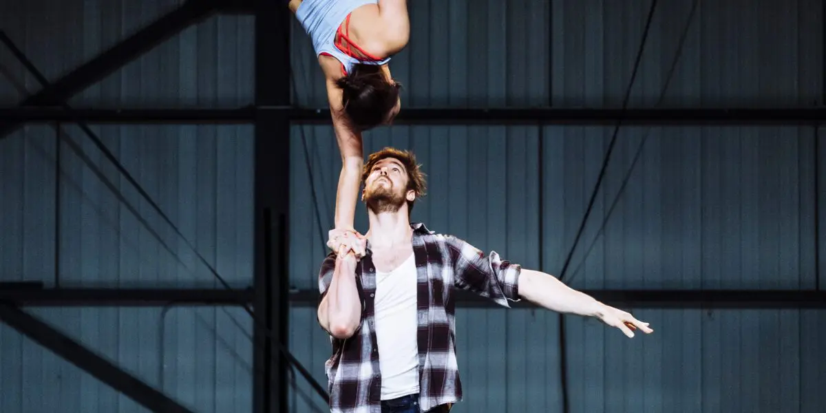Un homme fait tenir Ashley Youren en équilibre sur une main au-dessus de sa tête sur un tapis dans un espace ressemblant à un entrepôt. Elle a les jambes pliées et les bras tendus, tandis que l'homme est debout, pieds nus, un bras tendu.
