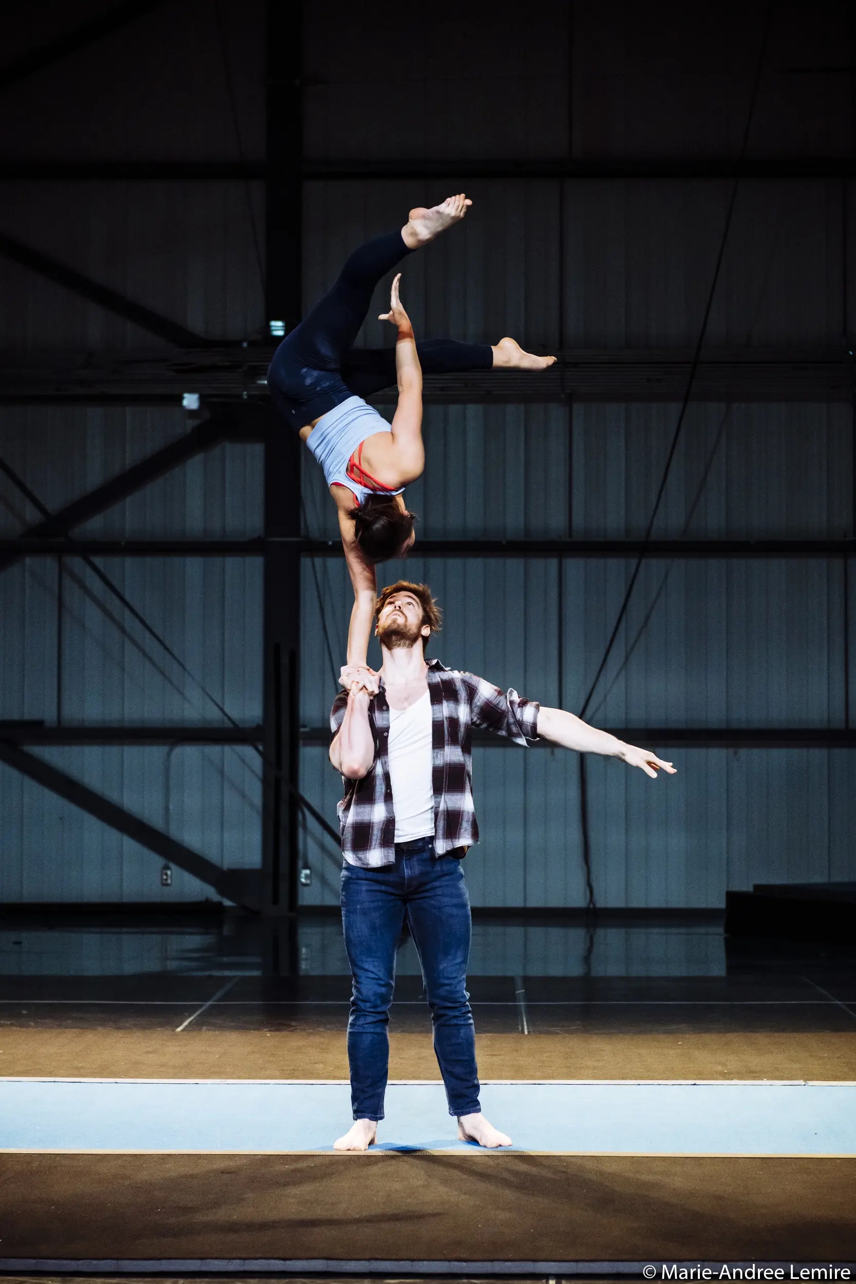 Un homme fait tenir Ashley Youren en équilibre sur une main au-dessus de sa tête sur un tapis dans un espace ressemblant à un entrepôt. Elle a les jambes pliées et les bras tendus, tandis que l'homme est debout, pieds nus, un bras tendu.