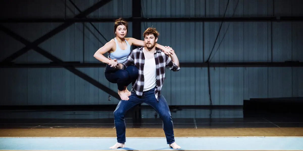 Ashley Youren se tient en équilibre, pieds nus et en tenue de sport, sur la jambe gauche pliée d'un homme qui se tient debout, en jean et chemise à carreaux, tous deux posés sur un tapis à l'intérieur d'une salle spacieuse de style industriel.
