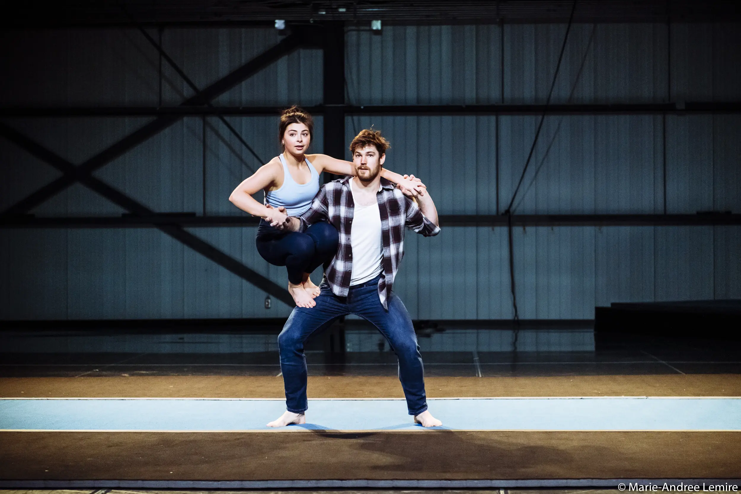 Ashley Youren se tient en équilibre, pieds nus et en tenue de sport, sur la jambe gauche pliée d'un homme qui se tient debout, en jean et chemise à carreaux, tous deux posés sur un tapis à l'intérieur d'une salle spacieuse de style industriel.