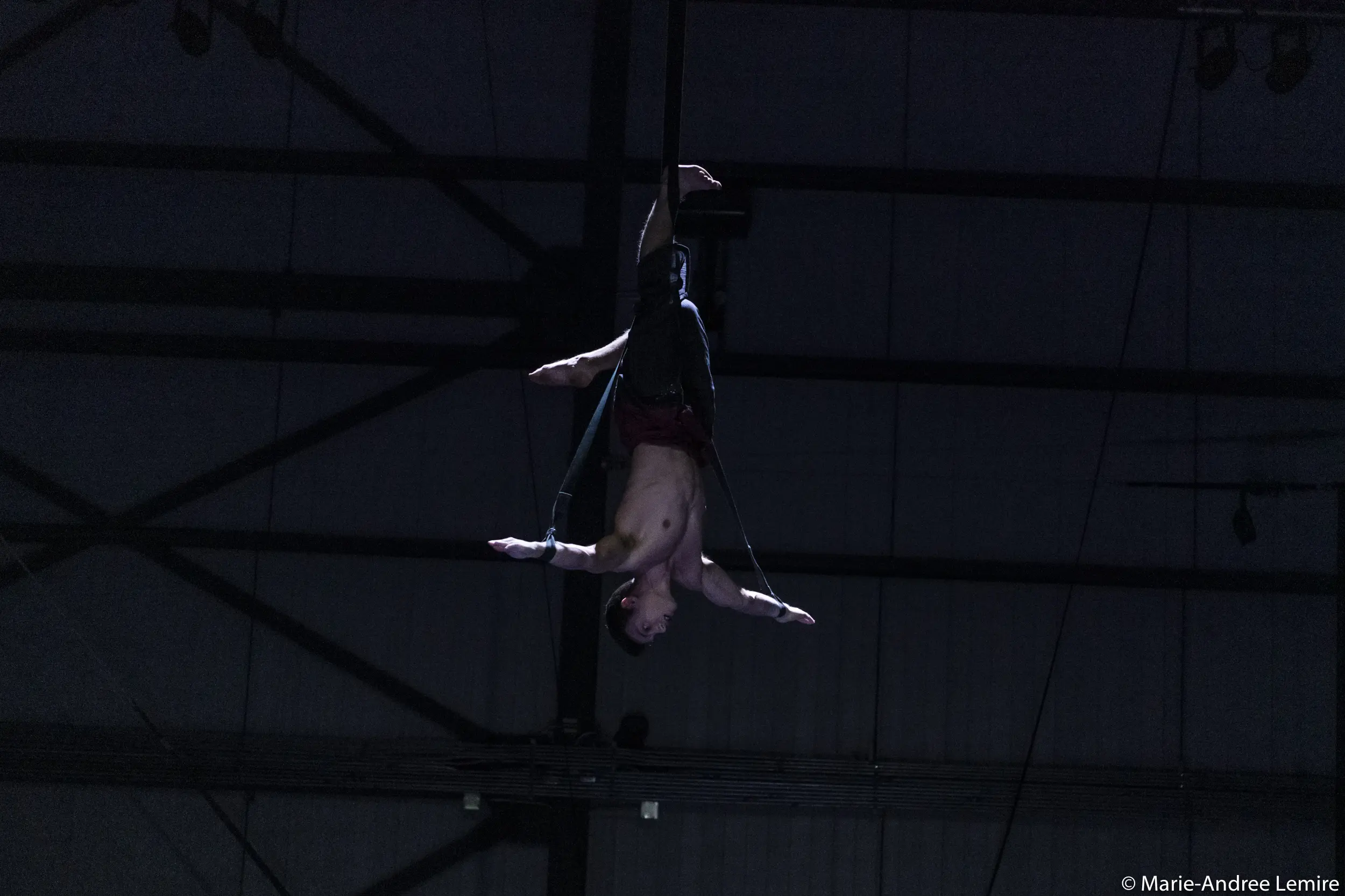 Un voltigeur, Samuel Donald Nash, est suspendu à des sangles, les bras étendus, sous un faible éclairage et dans un plafond industriel sombre.