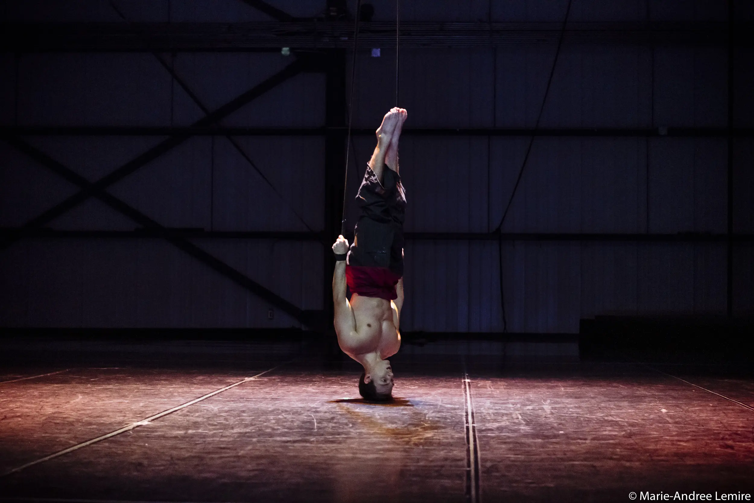 Un artiste torse nu, Samuel Donald Nash, est suspendu par les jambes au bout d'une corde dans un espace intérieur faiblement éclairé, avec un éclairage dramatique mettant en valeur son corps sur un fond industriel sombre.