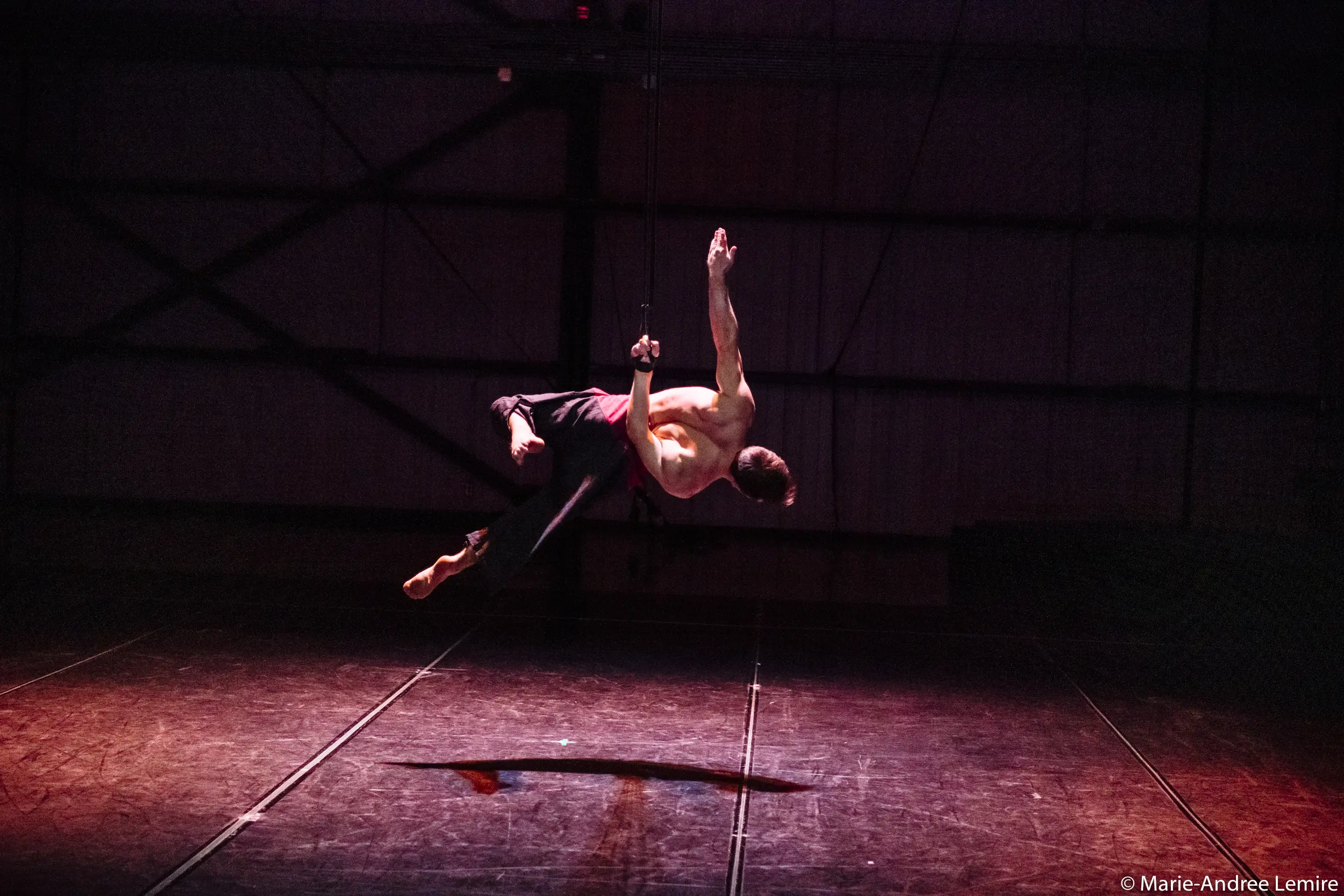 Un artiste aérien torse nu, Samuel Donald Nash, est gracieusement suspendu dans les airs sur un fond industriel faiblement éclairé, avec un éclairage dramatique qui projette des ombres sur le sol de la scène en contrebas.