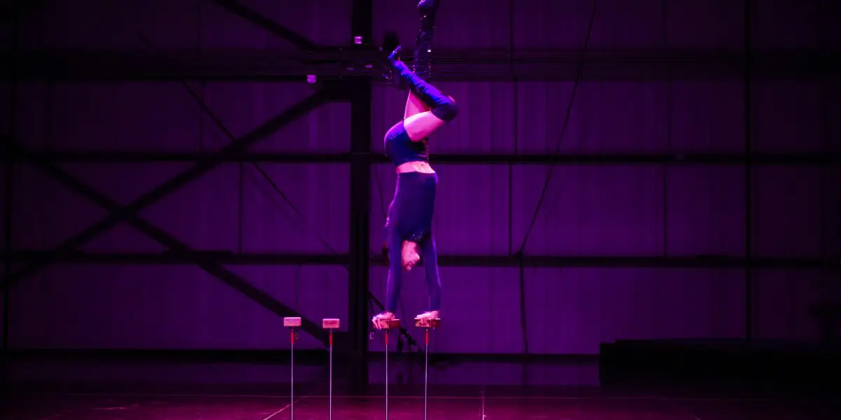 Un interprète vêtu de bottes et d'un costume noirs se balance à l'envers sur des cannes en équilibre sous un éclairage violet dramatique dans un grand espace intérieur de type industriel, évoquant le style de mise en scène novateur de Yann Lortie.