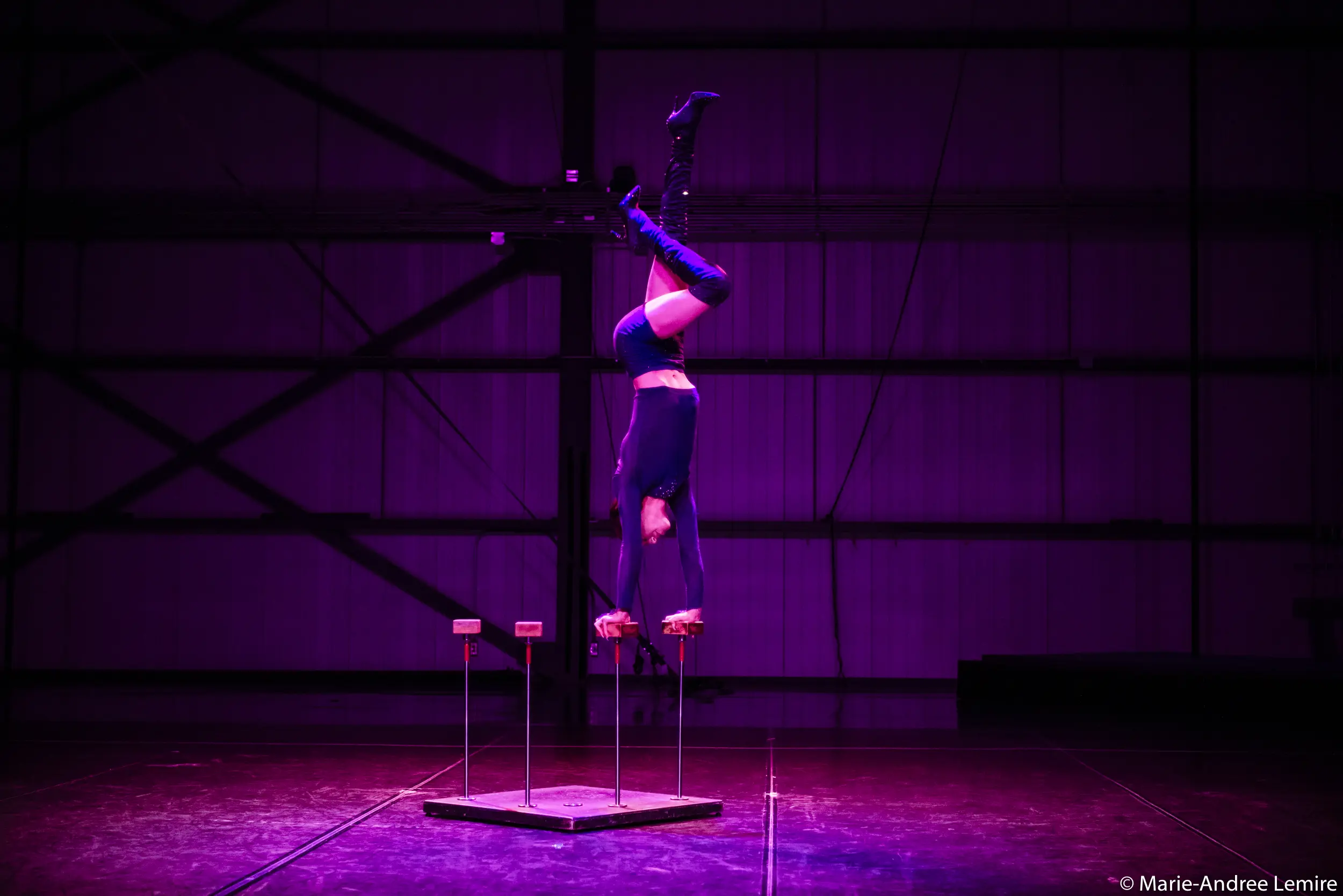 Un interprète vêtu de bottes et d'un costume noirs se balance à l'envers sur des cannes en équilibre sous un éclairage violet dramatique dans un grand espace intérieur de type industriel, évoquant le style de mise en scène novateur de Yann Lortie.
