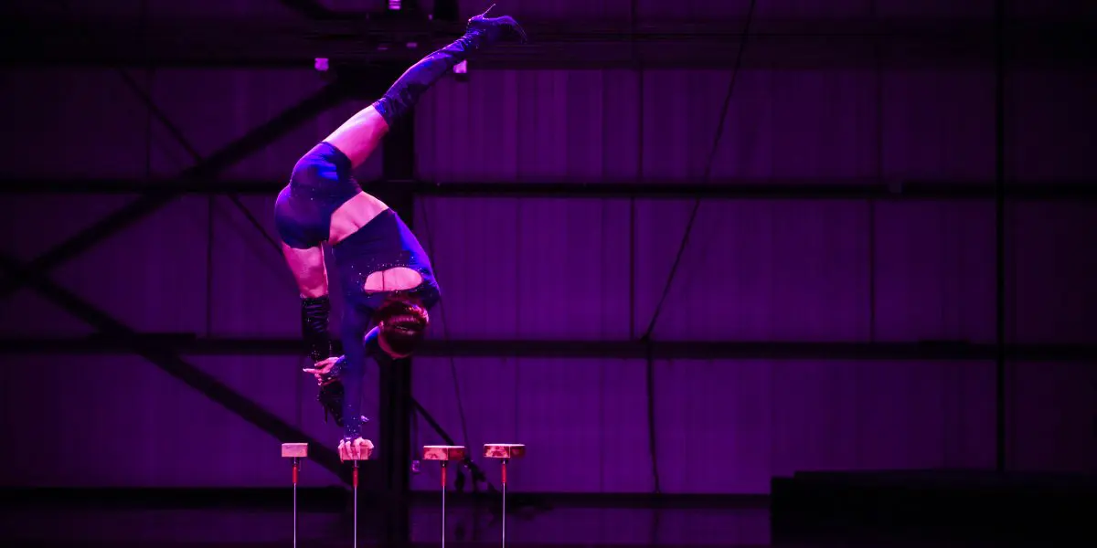 Un artiste de cirque en costume bleu, qui s'inspire de Yann Lortie, se tient en équilibre à l'envers sur une main au sommet de cannes, les jambes étendues en fente, sur un fond industriel éclairé en violet.