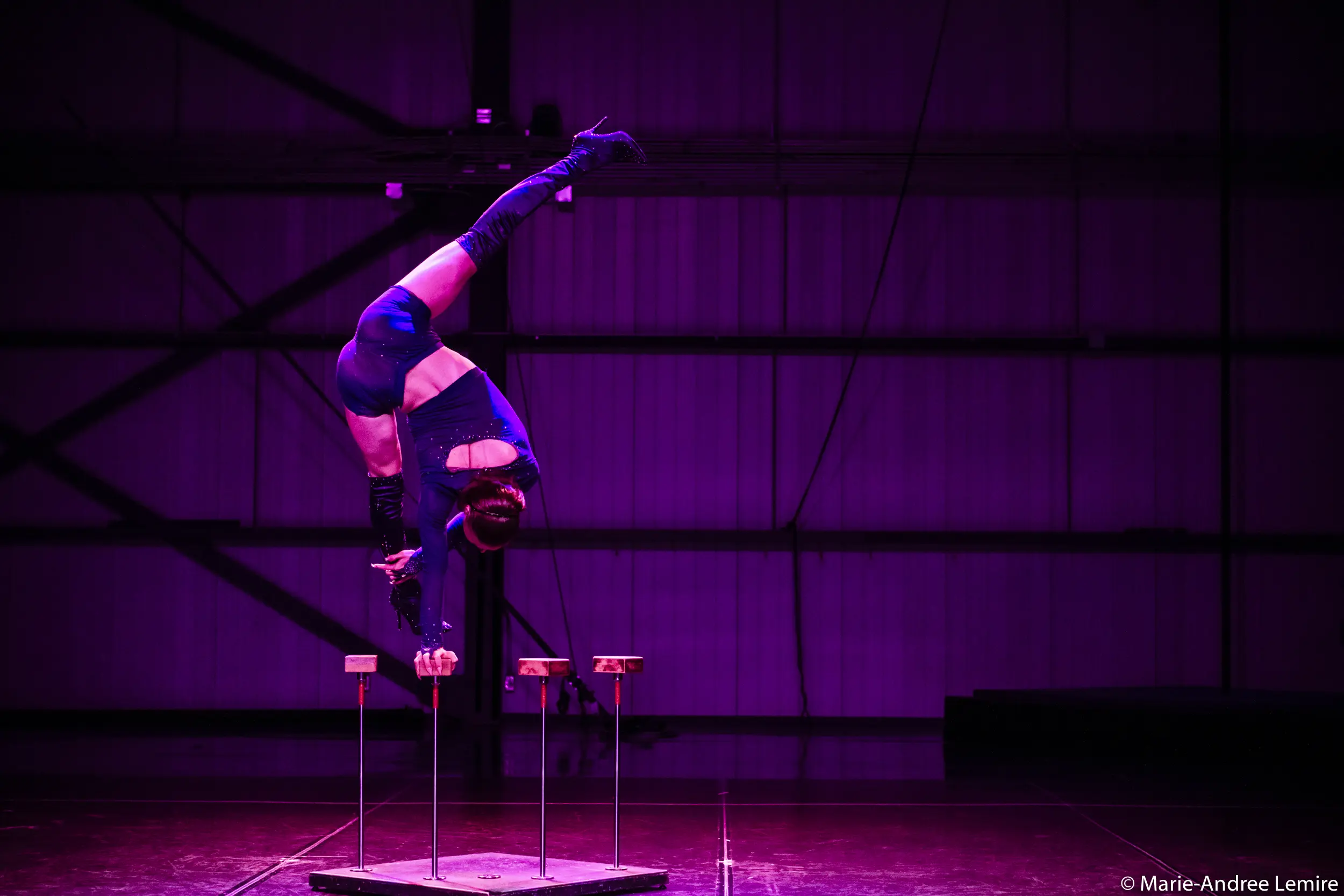 Un artiste de cirque en costume bleu, qui s'inspire de Yann Lortie, se tient en équilibre à l'envers sur une main au sommet de cannes, les jambes étendues en fente, sur un fond industriel éclairé en violet.