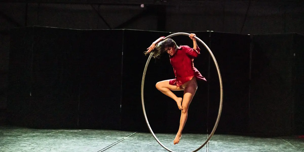 Andrea Martinez Aguilar, vêtue d'une chemise rouge et d'un short, se tient gracieusement en équilibre sur un grand cerceau métallique sur la scène, une jambe tendue et les bras en extension, sous un éclairage dramatique.