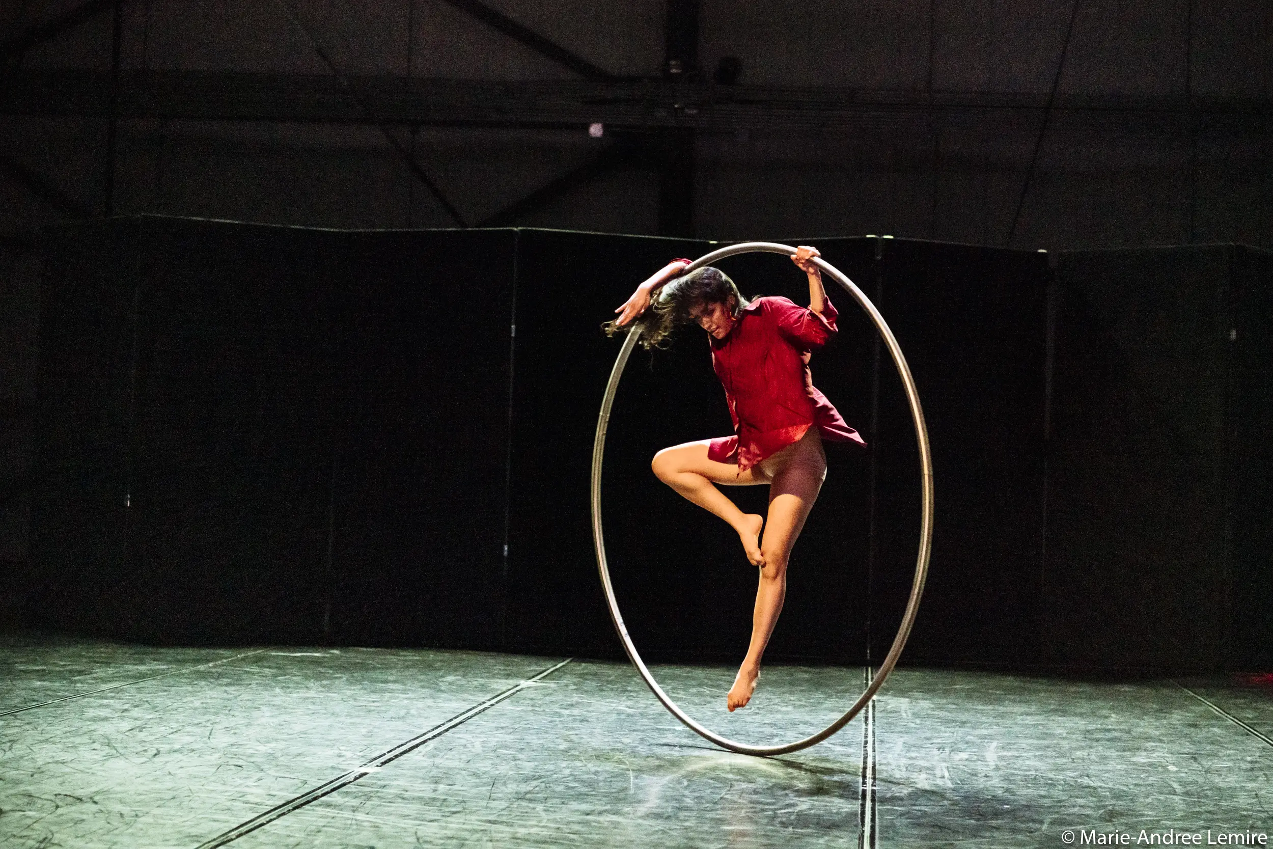 Andrea Martinez Aguilar, vêtue d'une chemise rouge et d'un short, se tient gracieusement en équilibre sur un grand cerceau métallique sur la scène, une jambe tendue et les bras en extension, sous un éclairage dramatique.