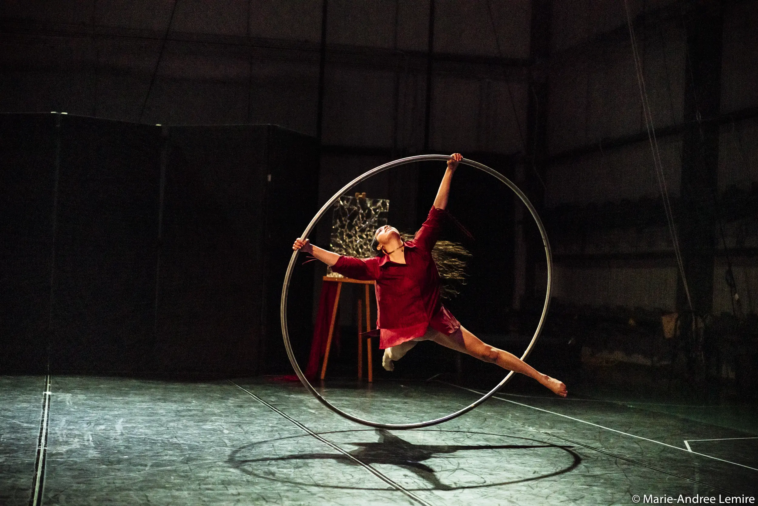 Andrea Martinez Aguilar, vêtue d'une tenue marron, se tient gracieusement en équilibre à l'intérieur d'un grand cerceau métallique sur une scène faiblement éclairée, un bras et une jambe tendus tandis qu'elle tourne. L'arrière-plan est sombre et les accessoires de scène sont visibles.