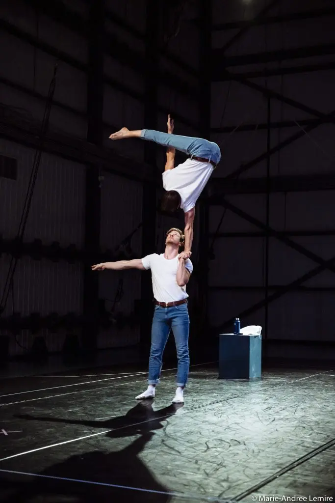 Un acrobate masculin, peut-être Benjamin Wolgers, tient en équilibre sur une main une partenaire féminine qui exécute un saut à l'envers ; tous deux portent des chemises blanches et des jeans bleus et se produisent sur une scène faiblement éclairée.