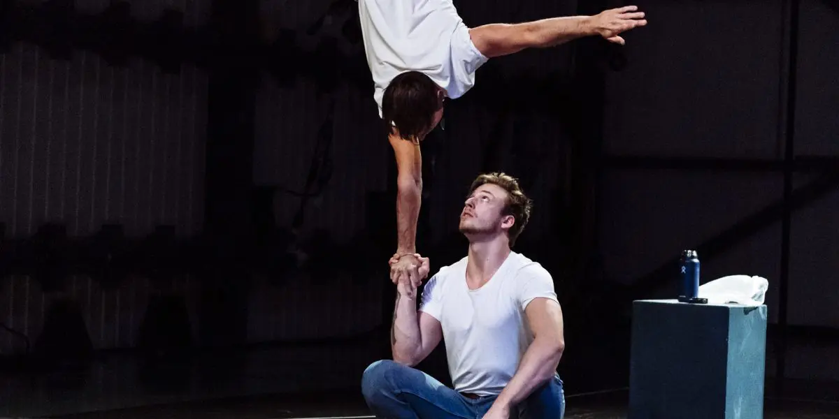 Deux acrobates, dont Benjamin Wolgers, se produisent à l'intérieur ; l'un se tient en équilibre sur une main au-dessus de la main de l'autre, tandis que le second acrobate s'agenouille pour soutenir son partenaire. Les deux acrobates portent des jeans et des chemises blanches.