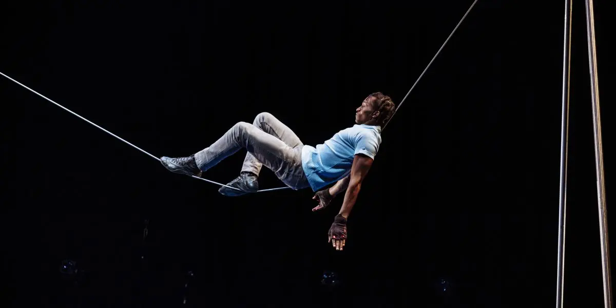 Antino Pansa, vêtu d'une chemise bleue et d'un pantalon gris, se tient habilement en équilibre sur une corde raide sur un fond sombre, une main touchant la corde et les jambes pliées pour se soutenir.