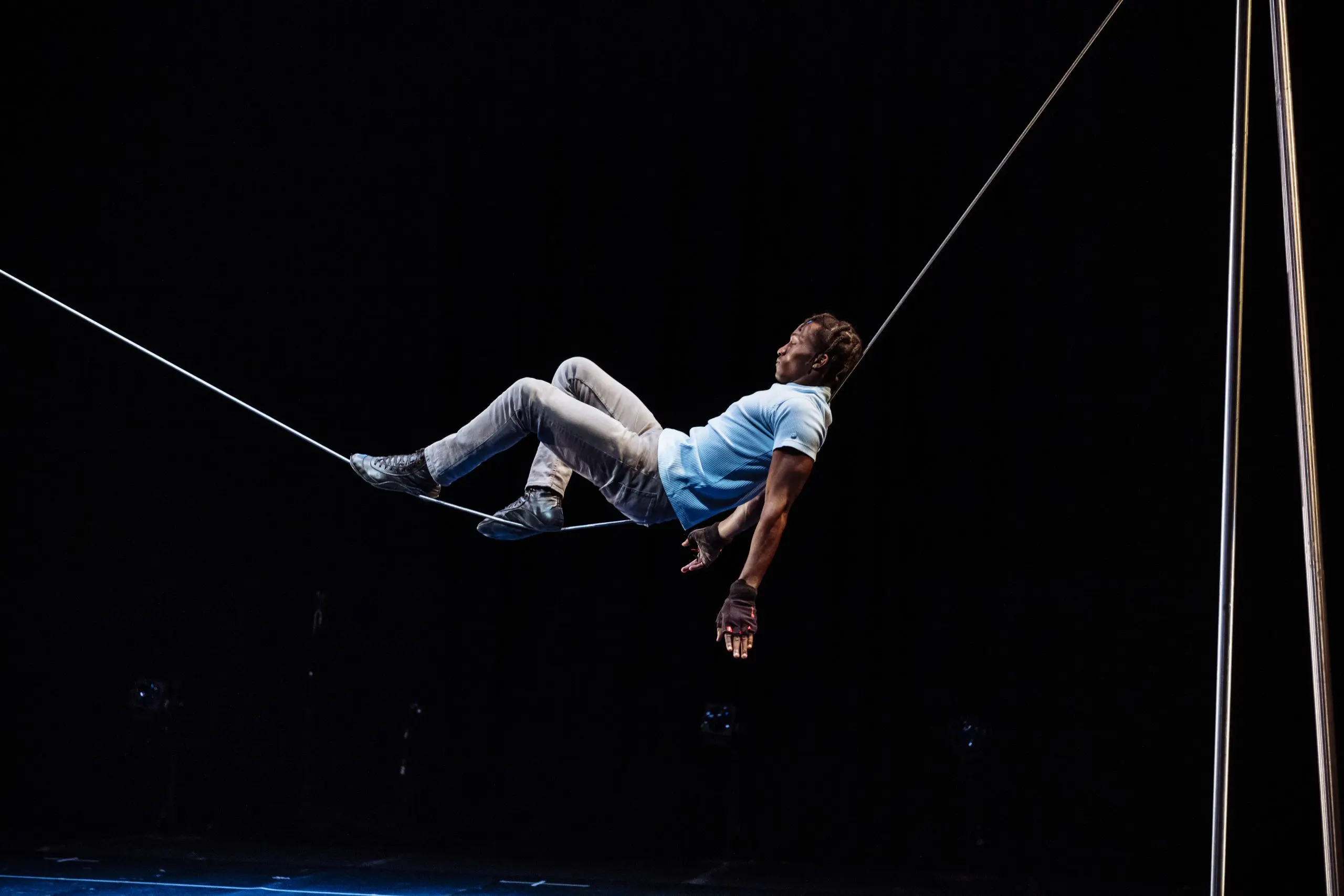 Antino Pansa, vêtu d'une chemise bleue et d'un pantalon gris, se tient habilement en équilibre sur une corde raide sur un fond sombre, une main touchant la corde et les jambes pliées pour se soutenir.