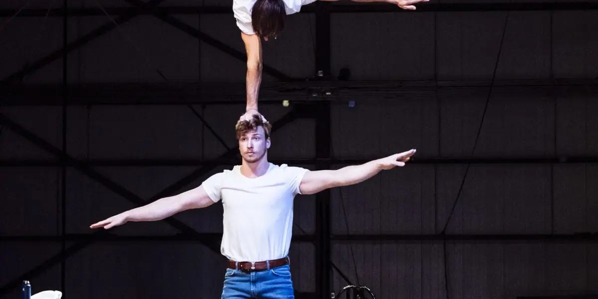 Deux artistes, dont Benjamin Wolgers, vêtus de chemises blanches et de jeans, se tiennent en équilibre sur la scène ; l'un se tient debout, les bras tendus, tandis que l'autre tient un poirier sur la tête, les jambes tendues en fente. Une chaise et une serviette sont à l'arrière-plan.