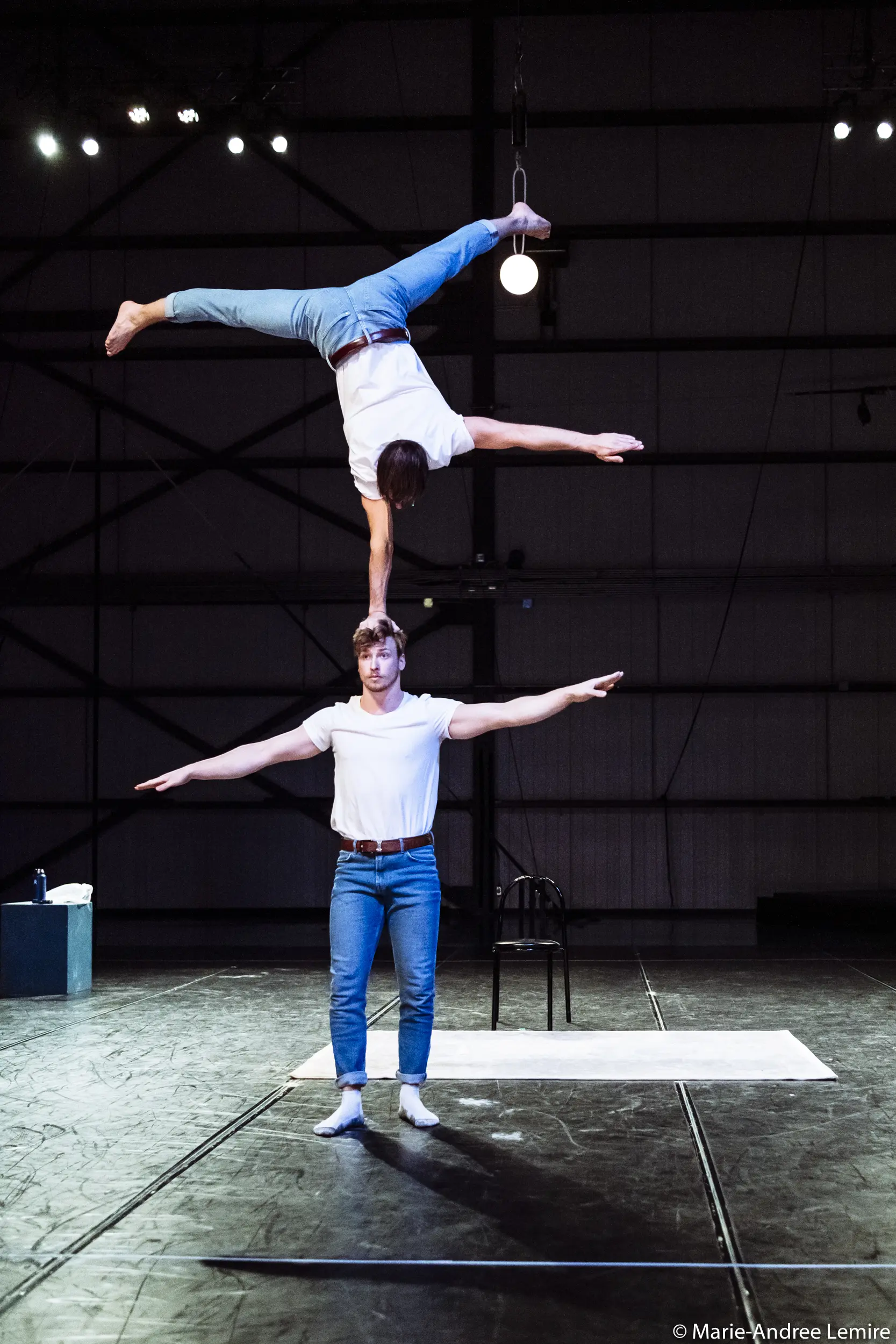 Deux artistes, dont Benjamin Wolgers, vêtus de chemises blanches et de jeans, se tiennent en équilibre sur la scène ; l'un se tient debout, les bras tendus, tandis que l'autre tient un poirier sur la tête, les jambes tendues en fente. Une chaise et une serviette sont à l'arrière-plan.