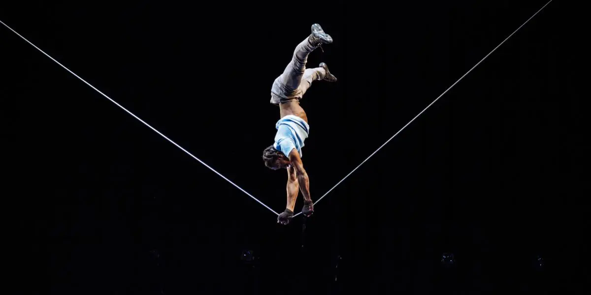 Antino Pansa, un artiste talentueux, se tient en équilibre sur une corde raide, la tête en bas, en s'agrippant à la corde avec les deux mains et les jambes tendues vers le haut, sur une scène au fond sombre.