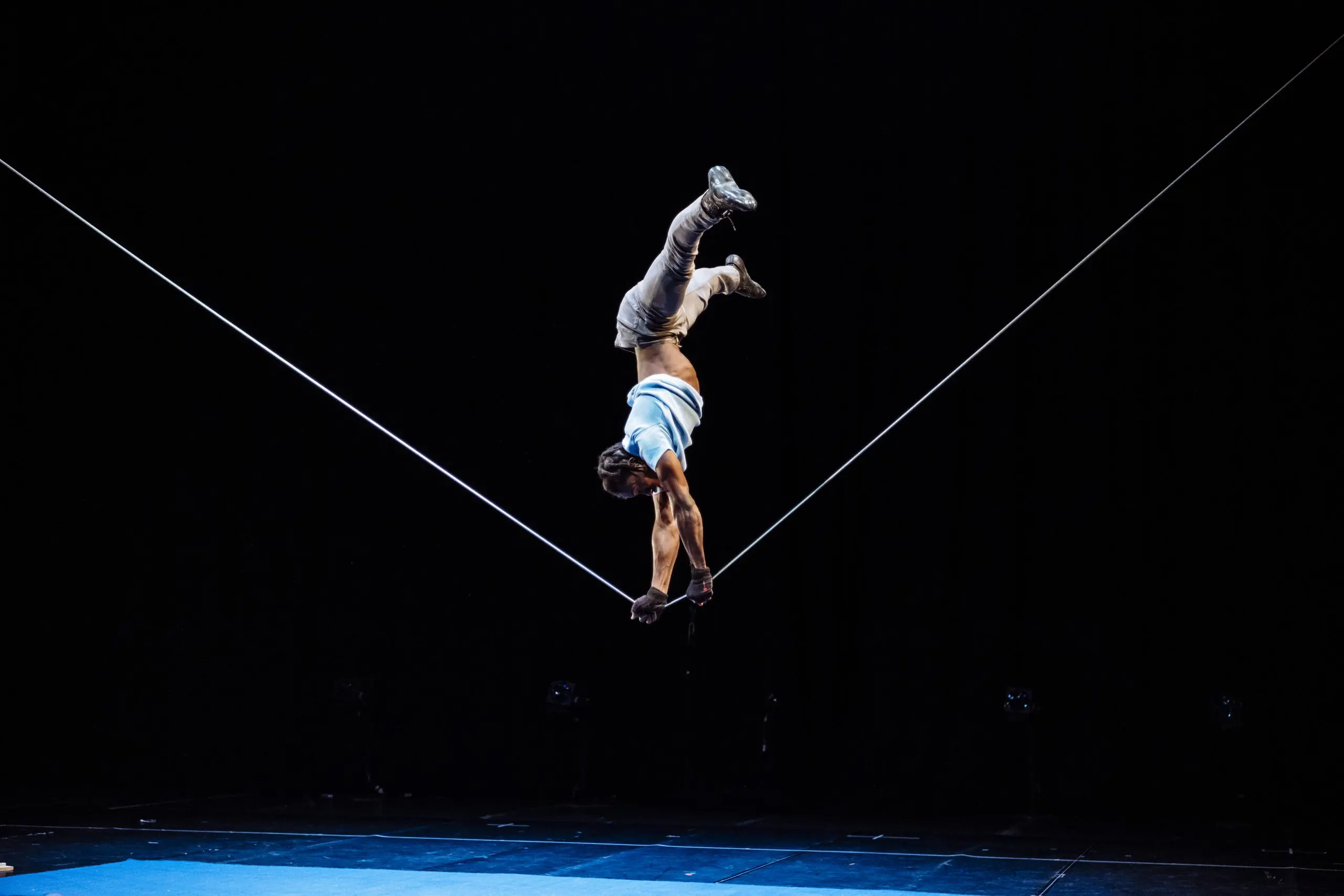 Antino Pansa, un artiste talentueux, se tient en équilibre sur une corde raide, la tête en bas, en s'agrippant à la corde avec les deux mains et les jambes tendues vers le haut, sur une scène au fond sombre.