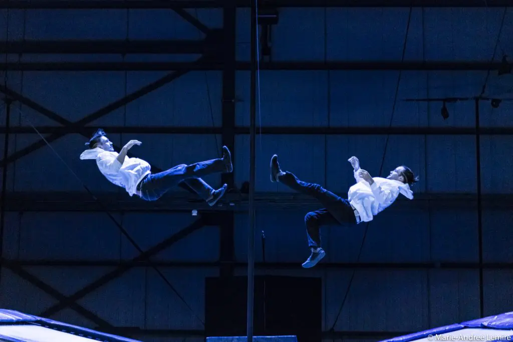Deux personnes vêtues de chemises blanches sont suspendues dans les airs, se faisant face et semblant tomber en arrière dans un espace intérieur faiblement éclairé, aux tons bleus, avec des poutres visibles et un grand poteau vertical entre elles - une image de Mathys Guyon Matte.