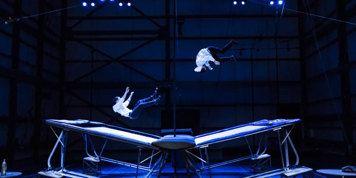 Deux performeurs en chemise blanche, dont Mathys Guyon Matte, sautent en l'air au-dessus de grands trampolines sur une scène faiblement éclairée par des projecteurs bleus dans un décor industriel sombre.