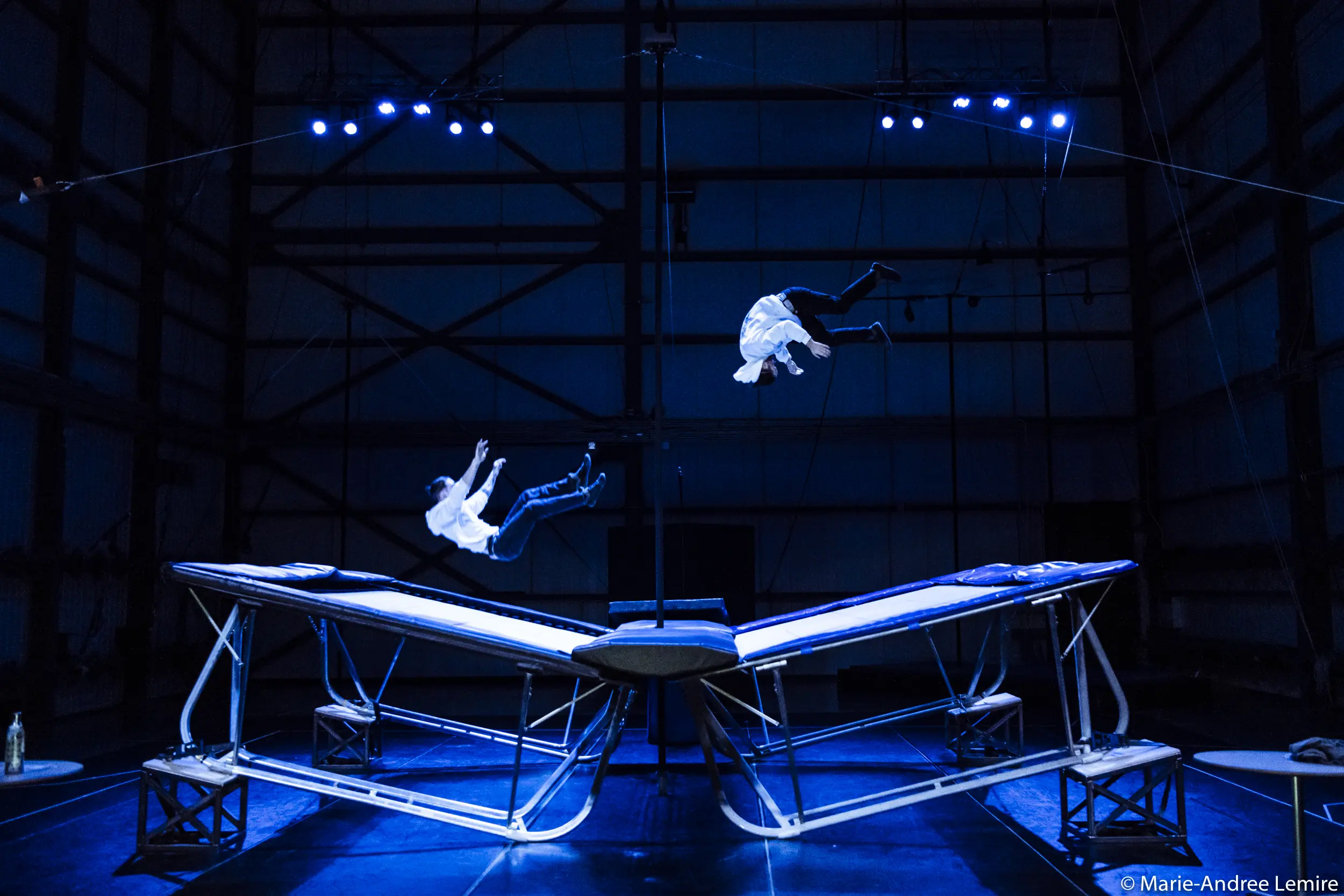 Deux performeurs en chemise blanche, dont Mathys Guyon Matte, sautent en l'air au-dessus de grands trampolines sur une scène faiblement éclairée par des projecteurs bleus dans un décor industriel sombre.