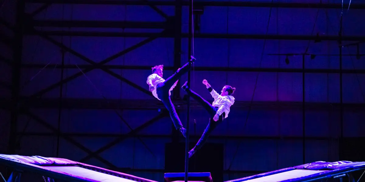 Deux acrobates, dont Mathys Guyon Matte, vêtus de chemises blanches et de pantalons sombres, exécutent une routine synchronisée en plein vol sur un trampoline, éclairés par un éclairage de scène bleu et rose spectaculaire, avec une grande structure métallique en arrière-plan.
