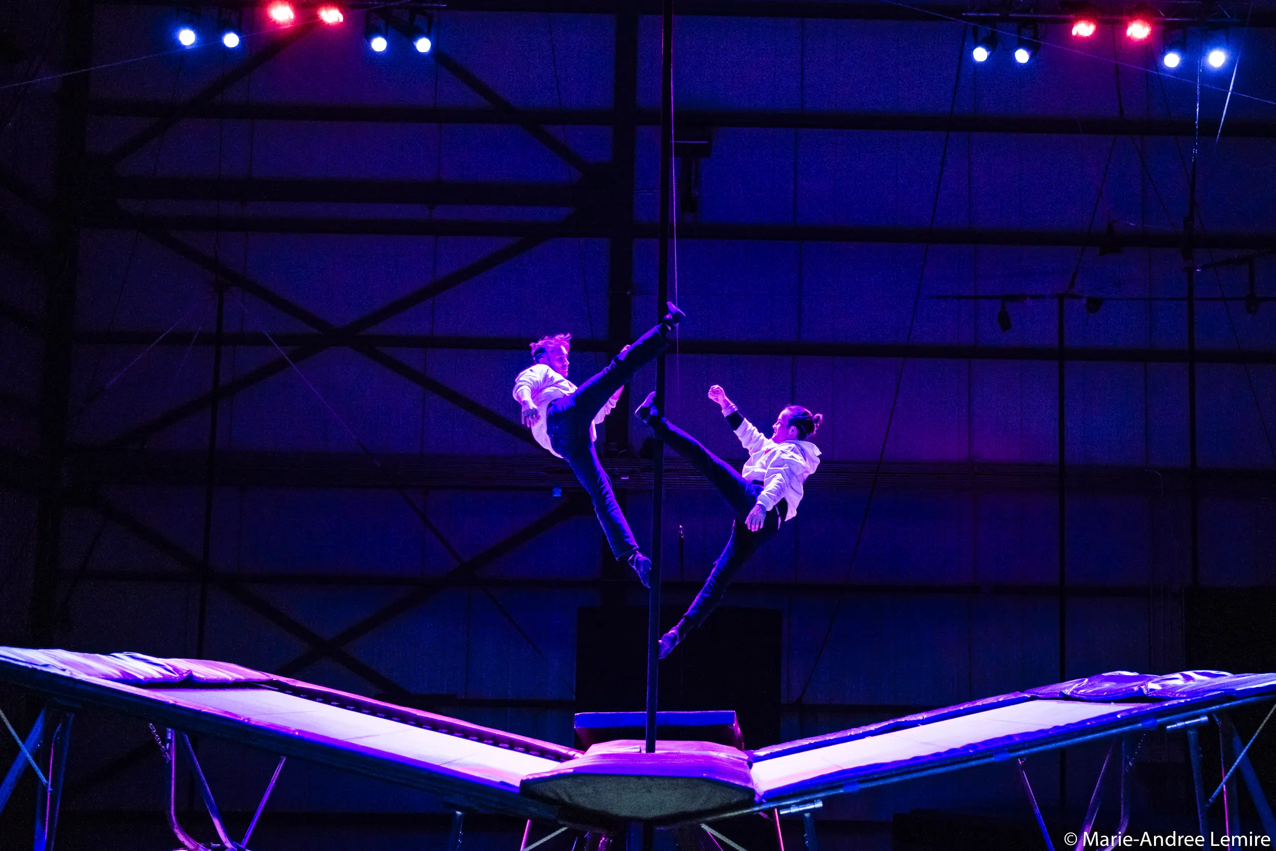 Deux acrobates, dont Mathys Guyon Matte, vêtus de chemises blanches et de pantalons sombres, exécutent une routine synchronisée en plein vol sur un trampoline, éclairés par un éclairage de scène bleu et rose spectaculaire, avec une grande structure métallique en arrière-plan.