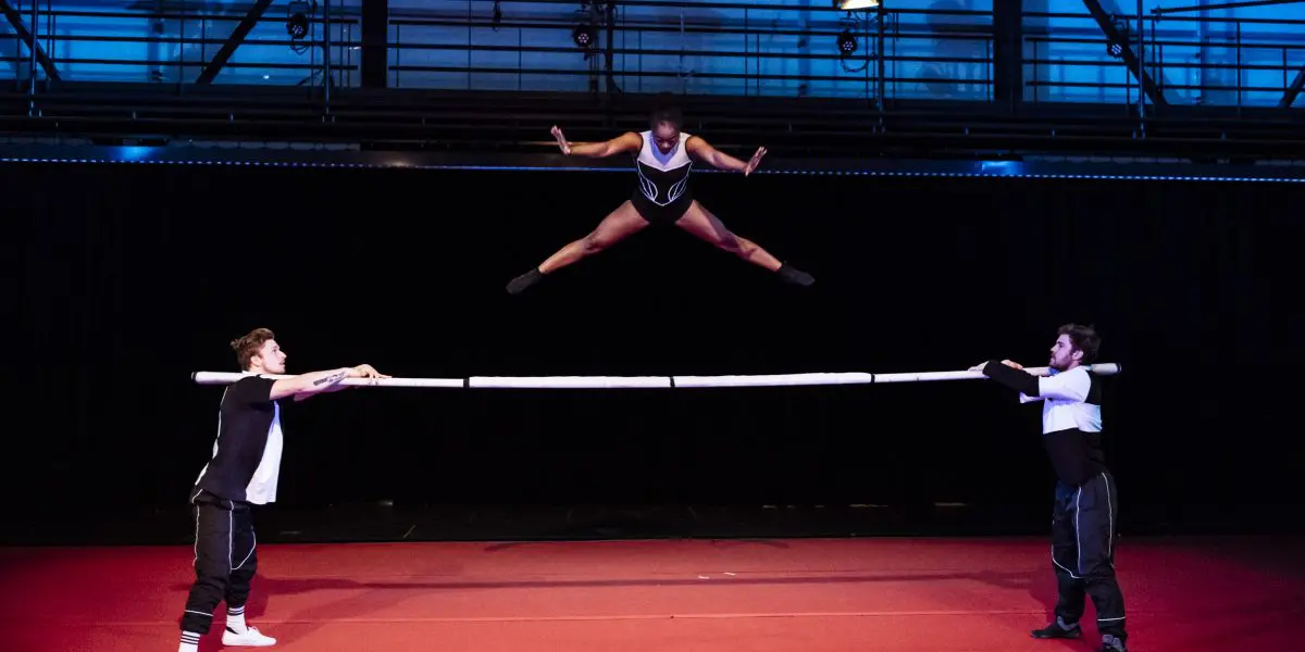 Naïka Aymon, une acrobate de talent, exécute un saut de puce en l'air au-dessus d'une barre horizontale tenue par deux artistes masculins sur un tapis rouge, avec un fond de scène sombre et un éclairage bleu au-dessus de la tête.