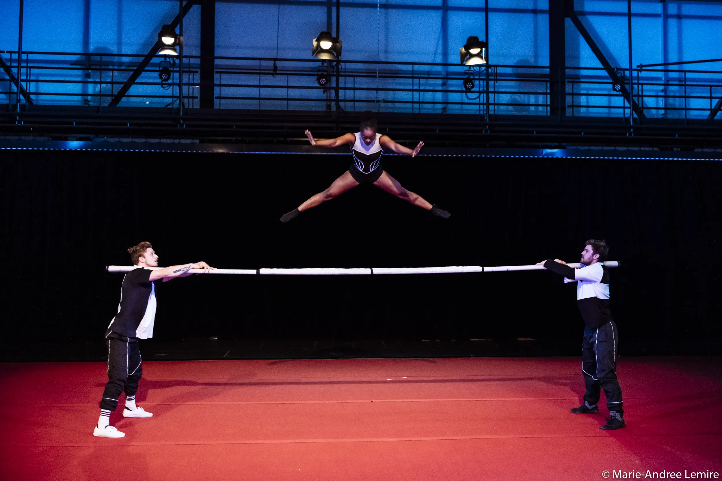 Naïka Aymon, une acrobate de talent, exécute un saut de puce en l'air au-dessus d'une barre horizontale tenue par deux artistes masculins sur un tapis rouge, avec un fond de scène sombre et un éclairage bleu au-dessus de la tête.