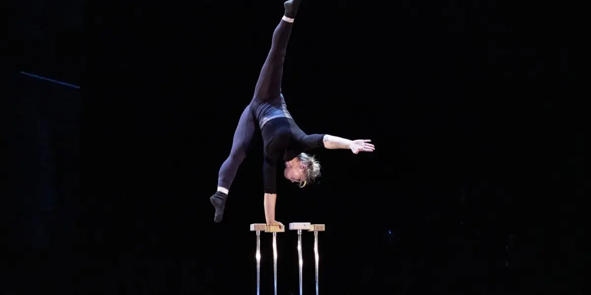 Amie Patching se tient en équilibre sur une main au sommet de deux cannes sur scène, avec une jambe tendue verticalement et l'autre inclinée, sur un fond sombre.