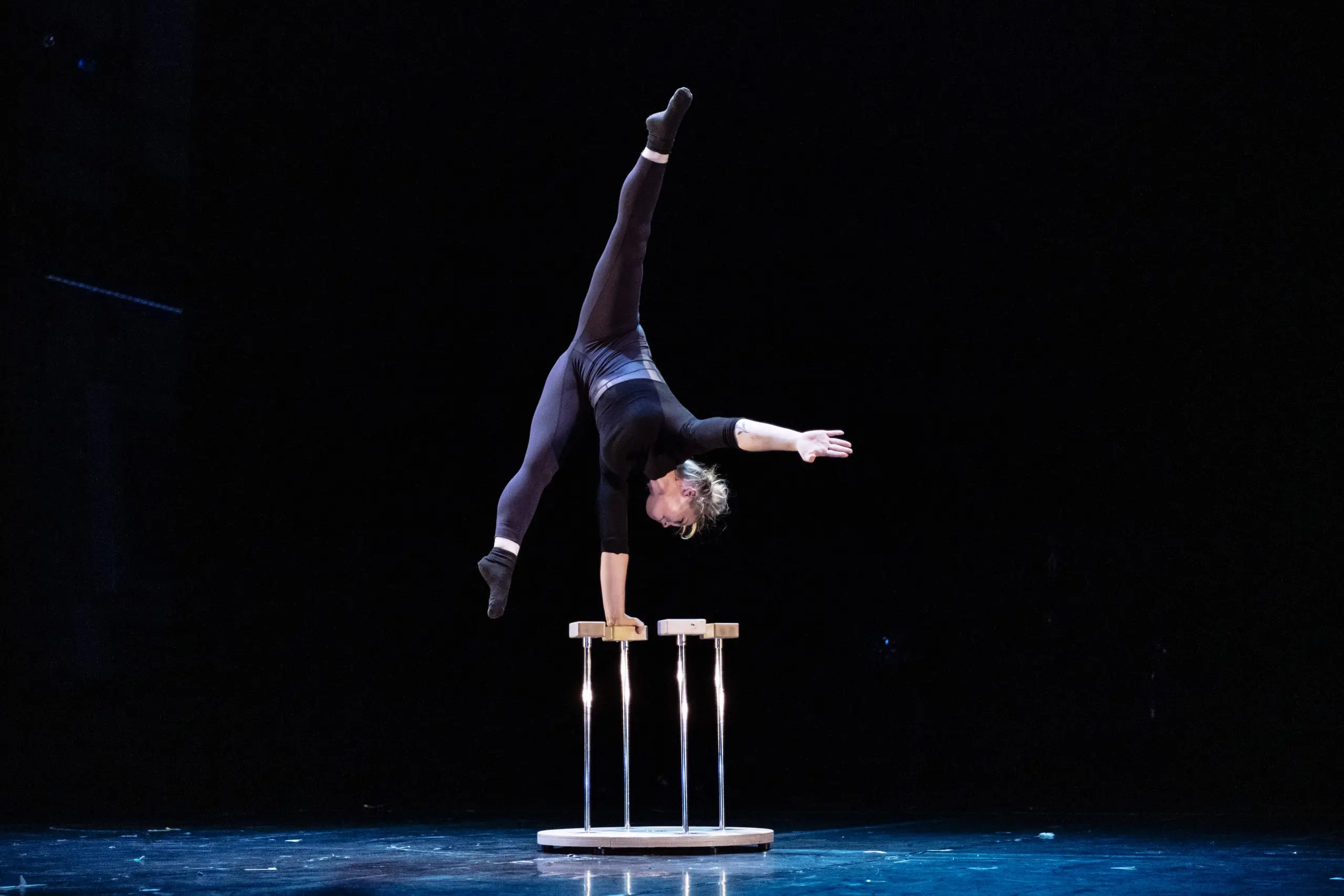 Amie Patching se tient en équilibre sur une main au sommet de deux cannes sur scène, avec une jambe tendue verticalement et l'autre inclinée, sur un fond sombre.