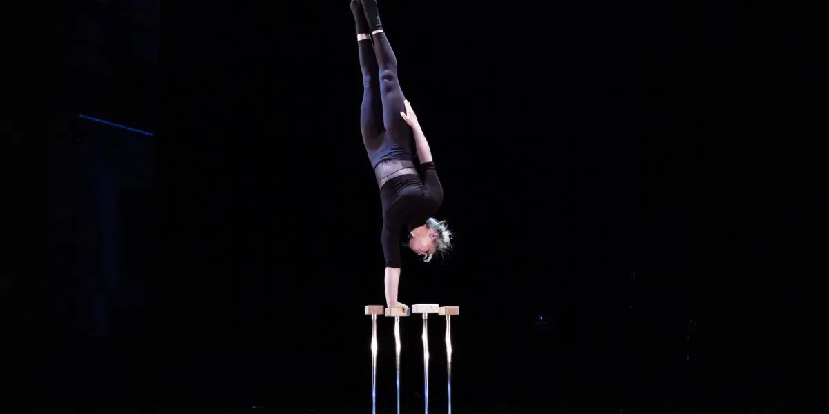 Amie Patching se tient en équilibre sur une main au sommet d'une paire de cannes sur une scène sombre, les jambes tendues et les orteils pointés, vêtue d'une tenue de sport sombre.