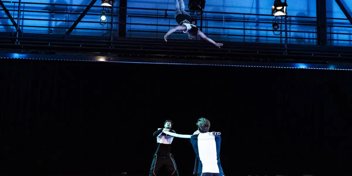 Trois artistes dans un numéro de cirque : deux se tiennent au sol en tenant des cordes tandis que Benjamin Wolgers est suspendu à l'envers dans les airs au-dessus d'eux, dans un décor intérieur dramatique et très éclairé.