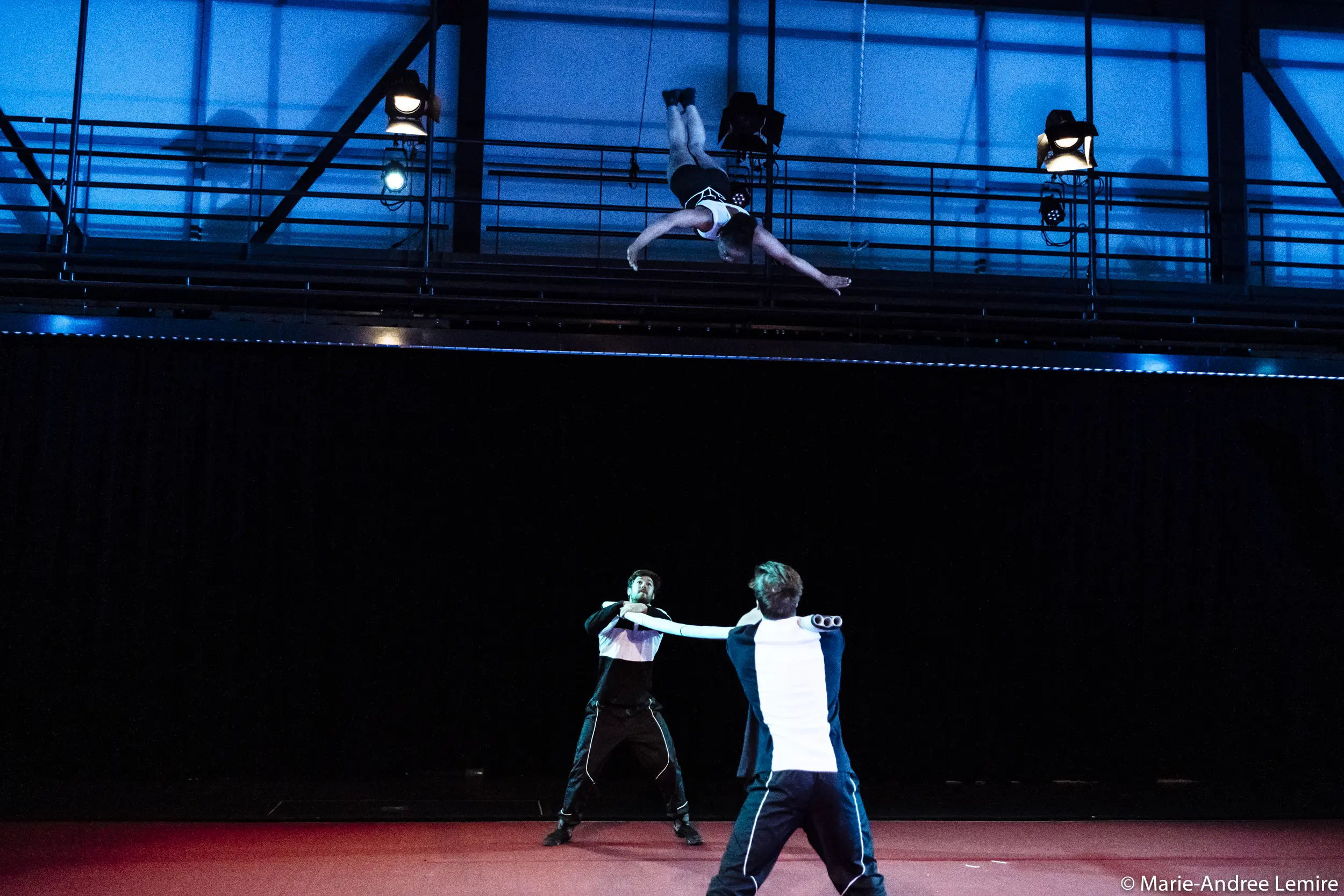 Trois artistes dans un numéro de cirque : deux se tiennent au sol en tenant des cordes tandis que Benjamin Wolgers est suspendu à l'envers dans les airs au-dessus d'eux, dans un décor intérieur dramatique et très éclairé.