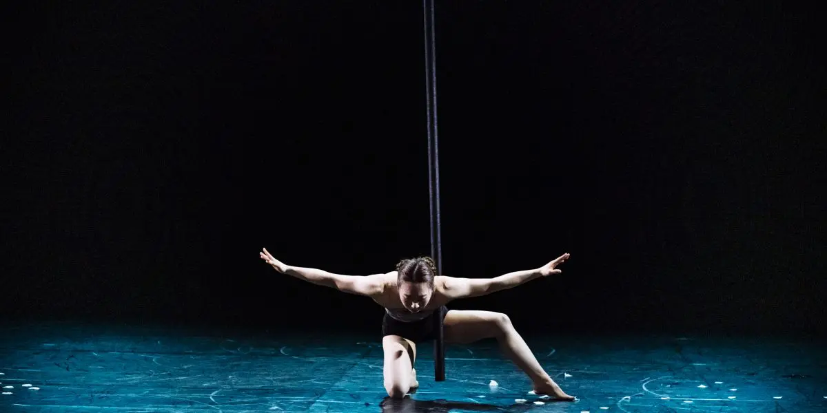 Danseuse en tenue noire, Minji Kim est agenouillée sur une jambe, les bras étendus, sous un poteau vertical, sur une scène faiblement éclairée au sol bleu.