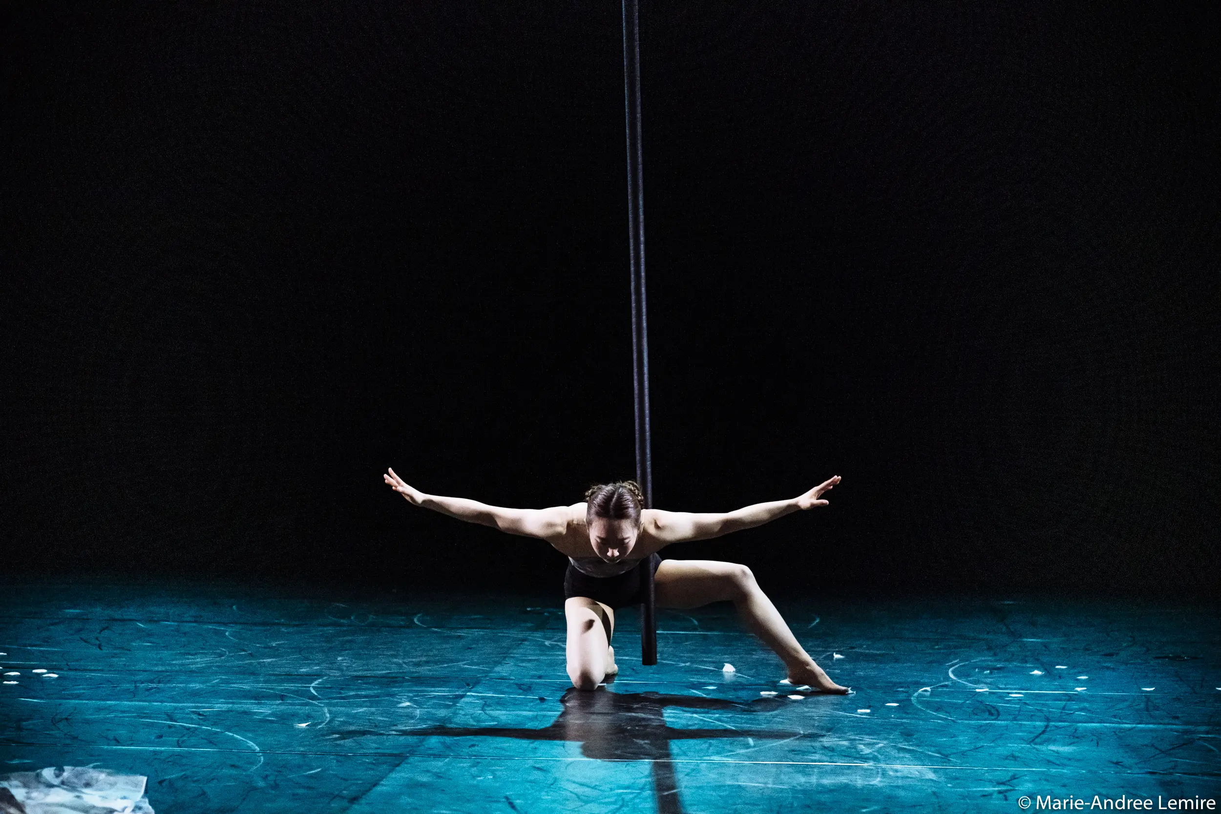 Danseuse en tenue noire, Minji Kim est agenouillée sur une jambe, les bras étendus, sous un poteau vertical, sur une scène faiblement éclairée au sol bleu.