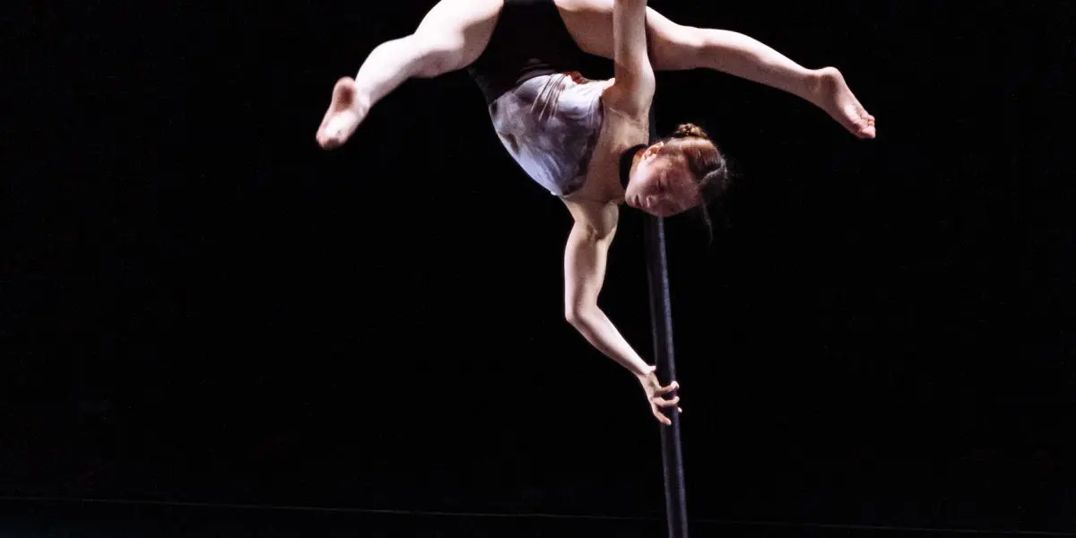 L'artiste aérienne Minji Kim, vêtue d'un haut blanc et d'un short noir, se suspend gracieusement à un poteau vertical, étendant ses jambes et un bras dans une pose dramatique sur une scène faiblement éclairée.