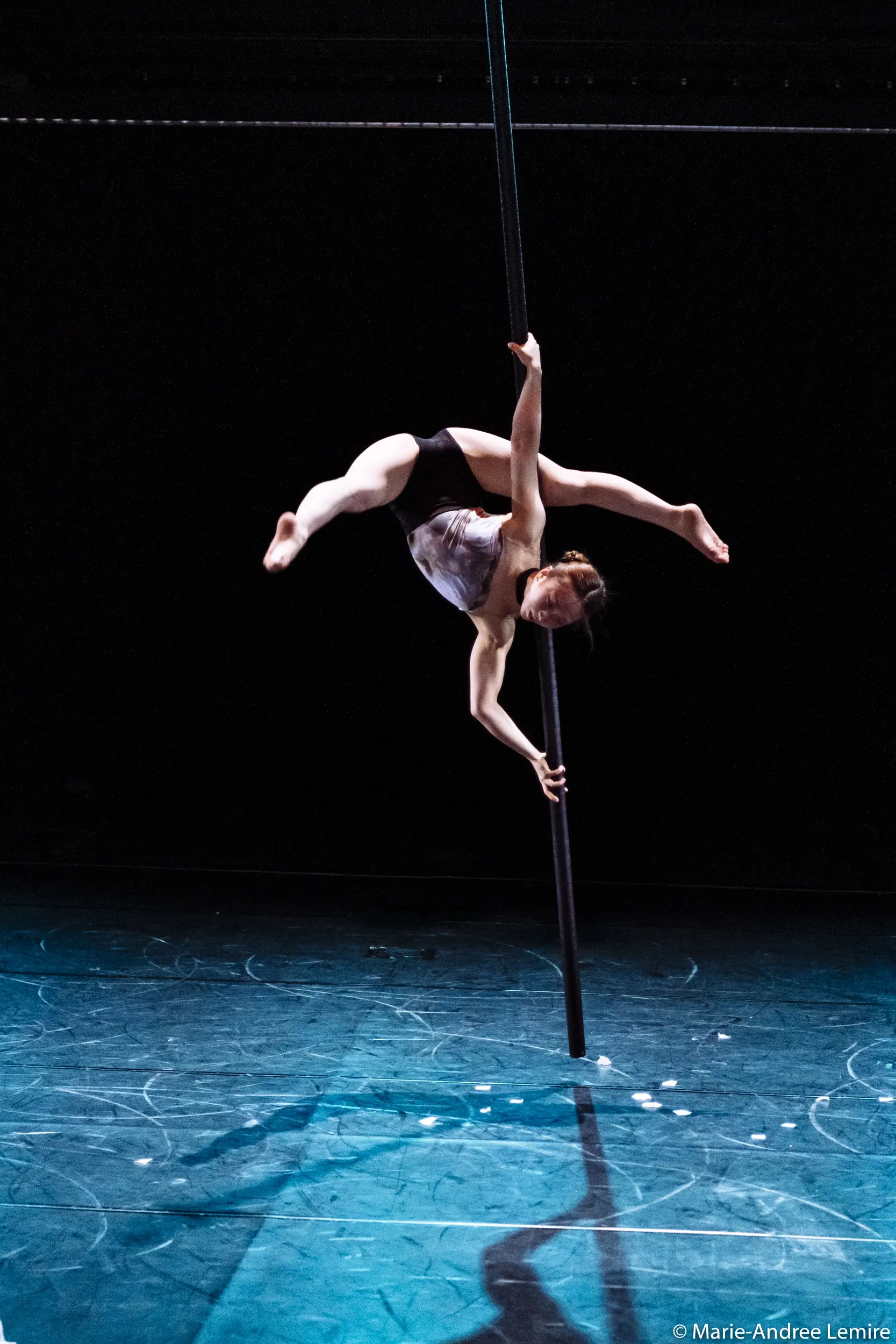L'artiste aérienne Minji Kim, vêtue d'un haut blanc et d'un short noir, se suspend gracieusement à un poteau vertical, étendant ses jambes et un bras dans une pose dramatique sur une scène faiblement éclairée.