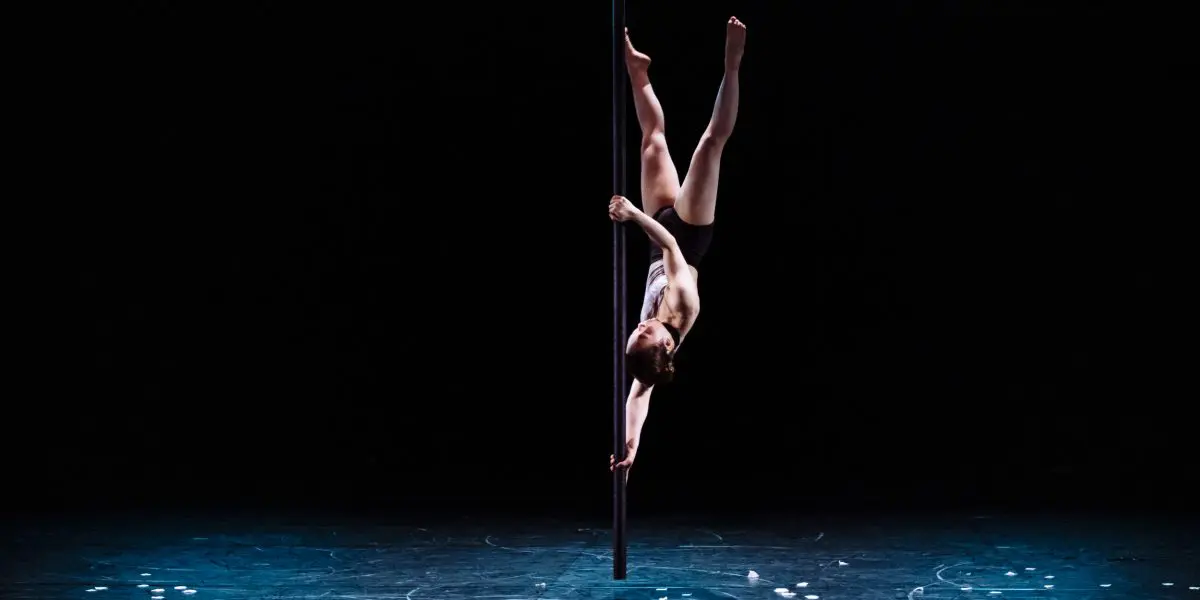 Minji Kim exécute une fente inversée, en saisissant la barre avec une jambe et un bras, sur une scène faiblement éclairée à l'arrière-plan sombre. Des objets blancs épars sont visibles sur le sol.