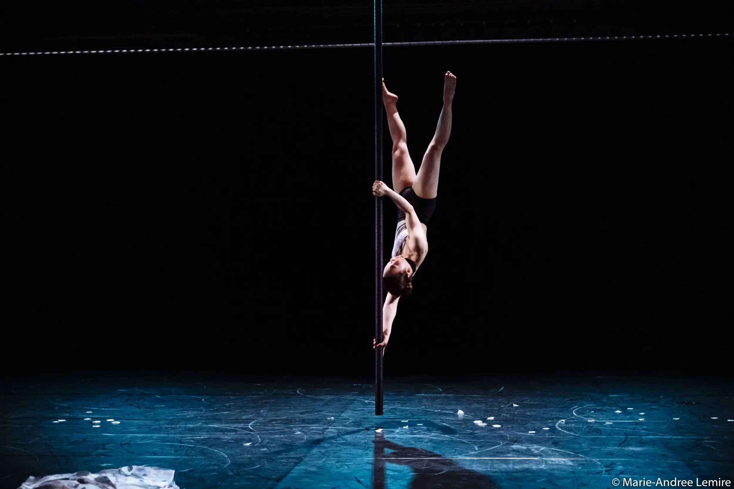 Minji Kim exécute une fente inversée, en saisissant la barre avec une jambe et un bras, sur une scène faiblement éclairée à l'arrière-plan sombre. Des objets blancs épars sont visibles sur le sol.