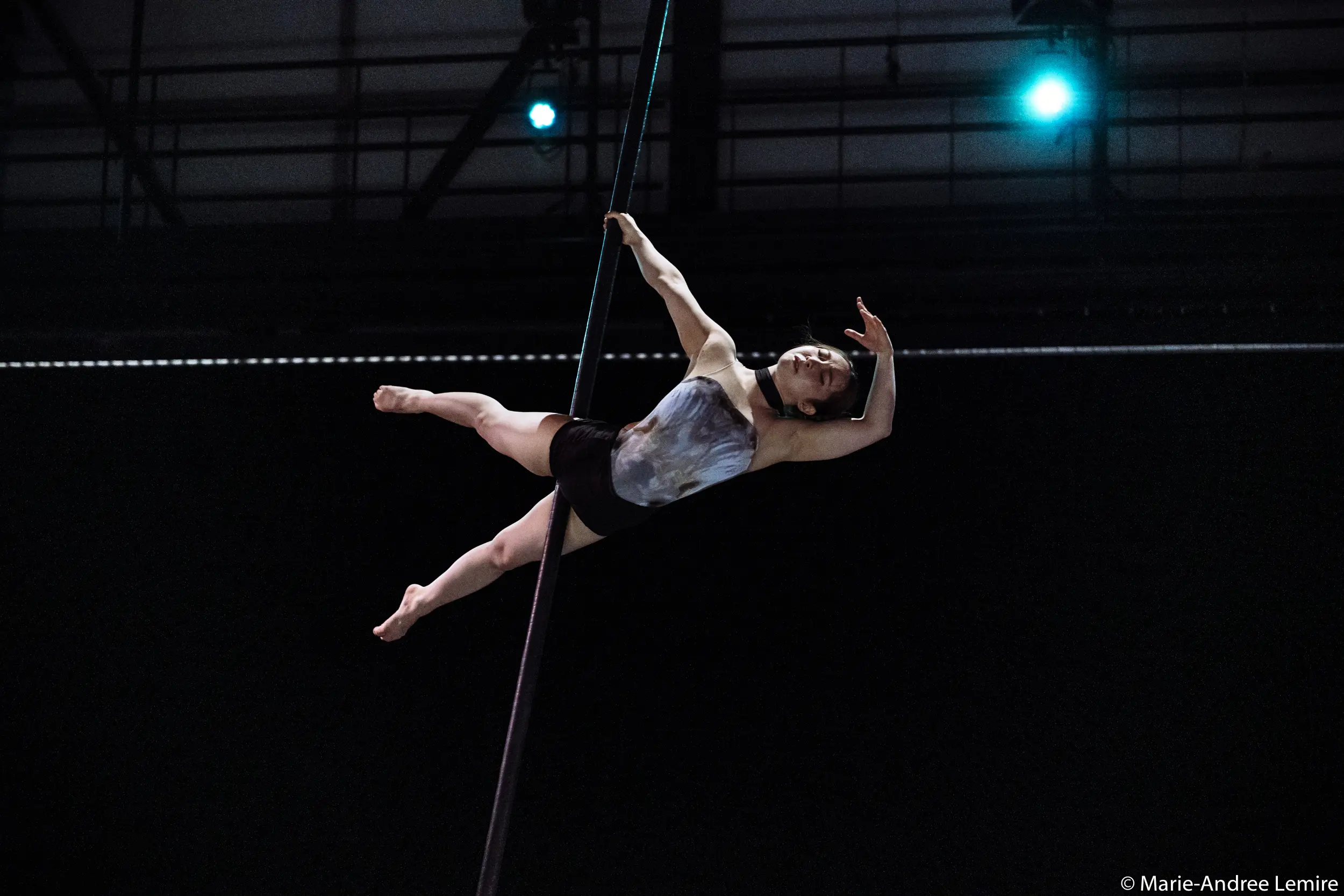 L'artiste aérienne Minji Kim prend une pose élégante alors qu'elle est suspendue horizontalement à une corde verticale, les lumières de la scène l'éclairant sur un fond sombre.