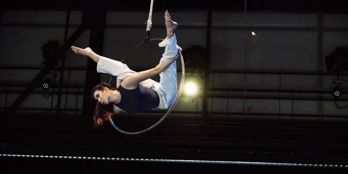 L'artiste aérienne Michelle Sargent se produit sur un cerceau suspendu, en équilibre avec une jambe crochetée et les bras étendus, sur un fond industriel. Elle porte un haut sombre et un pantalon clair.