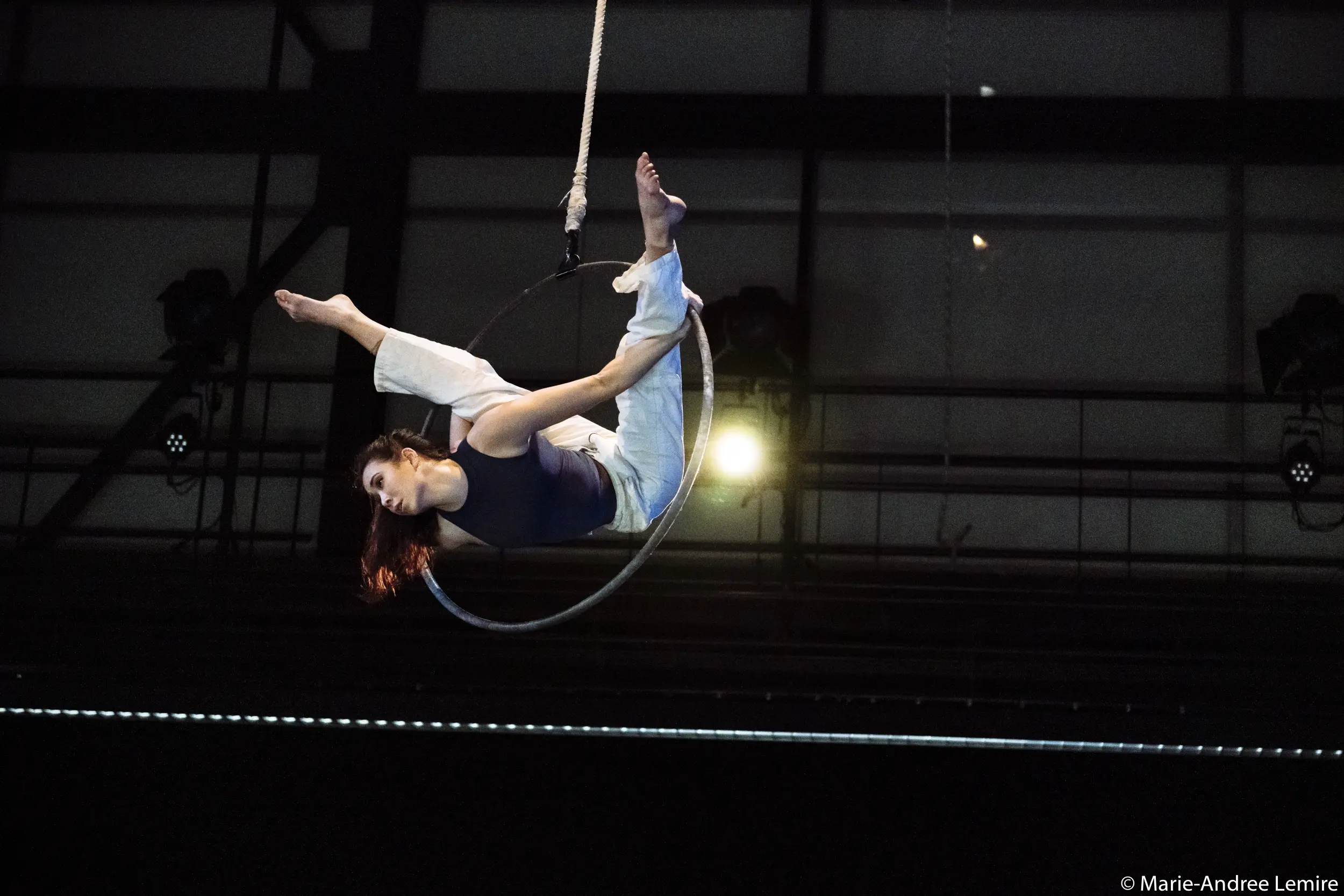 L'artiste aérienne Michelle Sargent se produit sur un cerceau suspendu, en équilibre avec une jambe crochetée et les bras étendus, sur un fond industriel. Elle porte un haut sombre et un pantalon clair.