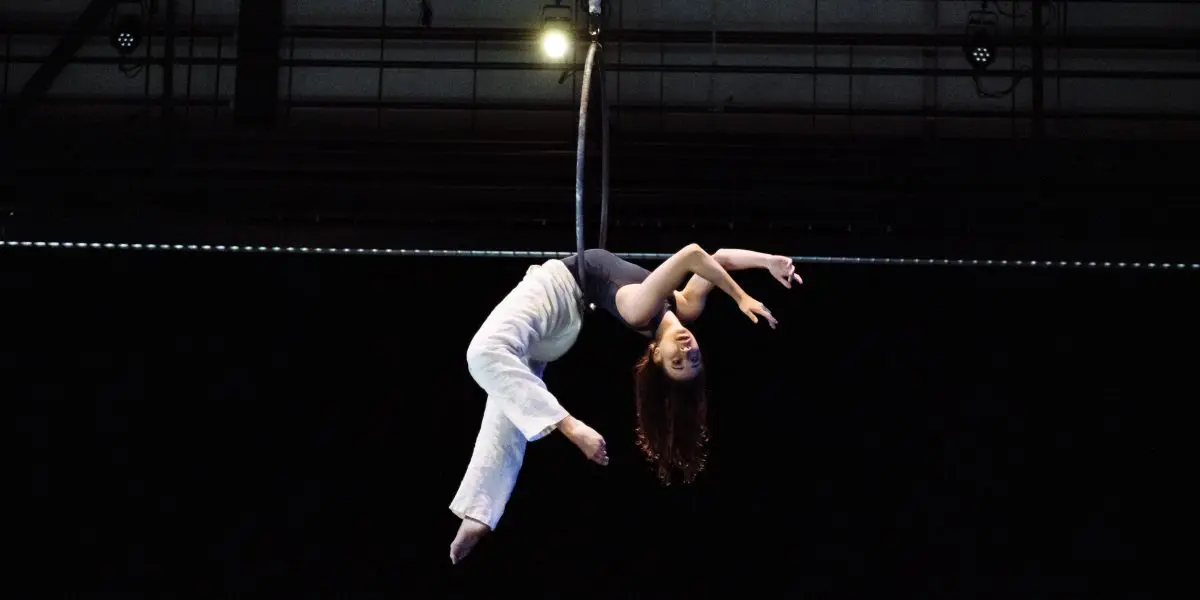 L'artiste aérienne Michelle Sargent est suspendue à l'envers sur un cerceau, vêtue d'un pantalon blanc et d'un haut gris, les bras gracieusement étendus sur un fond sombre.